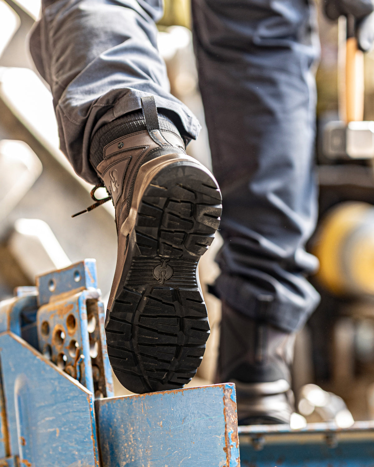 A close-up of a person's foot wearing a dark brown work boot with a black treaded sole, stepping onto a blue metal platform in an industrial setting.