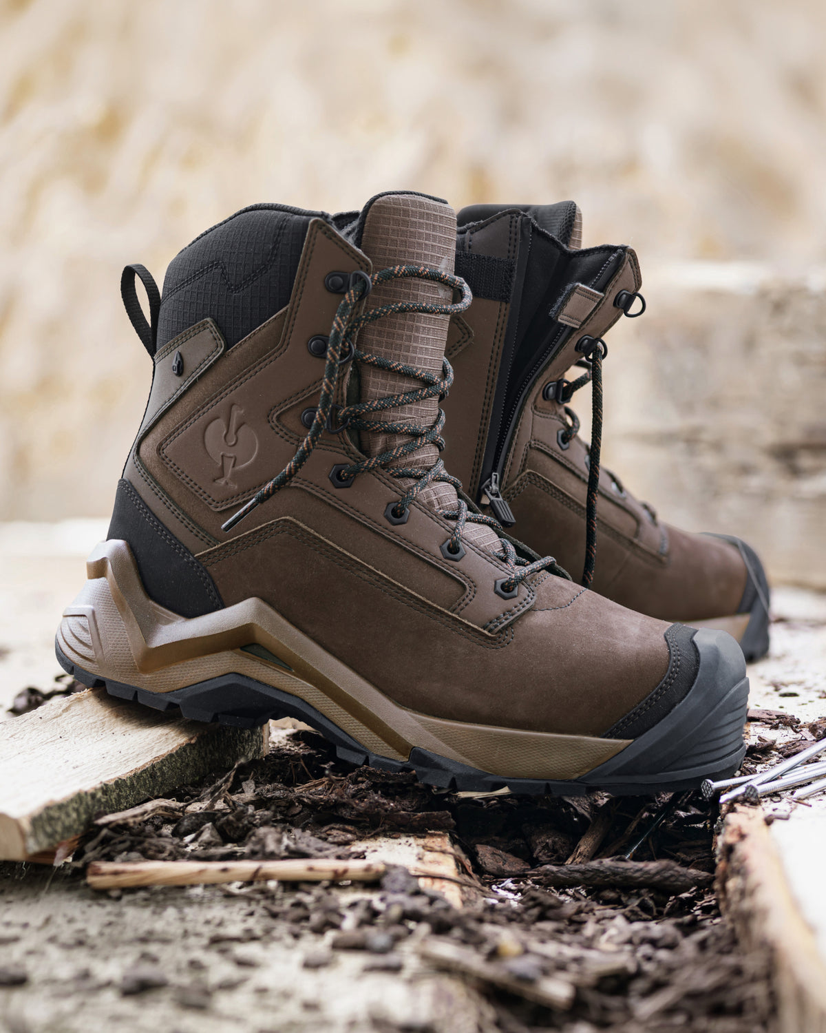 A pair of rugged brown tactical boots with black and tan accents, displayed on a rustic wooden surface with soil and twigs in the foreground.