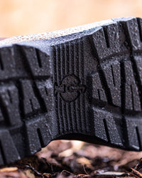 Close-up of a black rubber boot sole with a textured tread pattern and a circular logo embossed in the center, shown outdoors on a muddy surface with water droplets.
