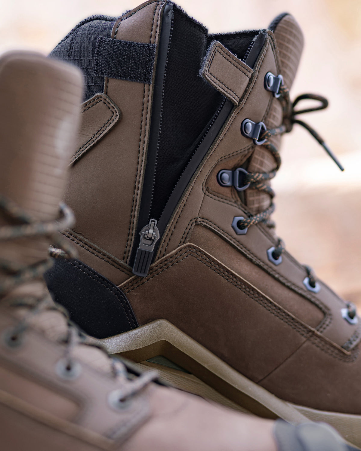 A close-up of a brown leather lace-up boot with a black side zipper, black ankle patch, and silver metal eyelets, shown against a blurred warm beige background.