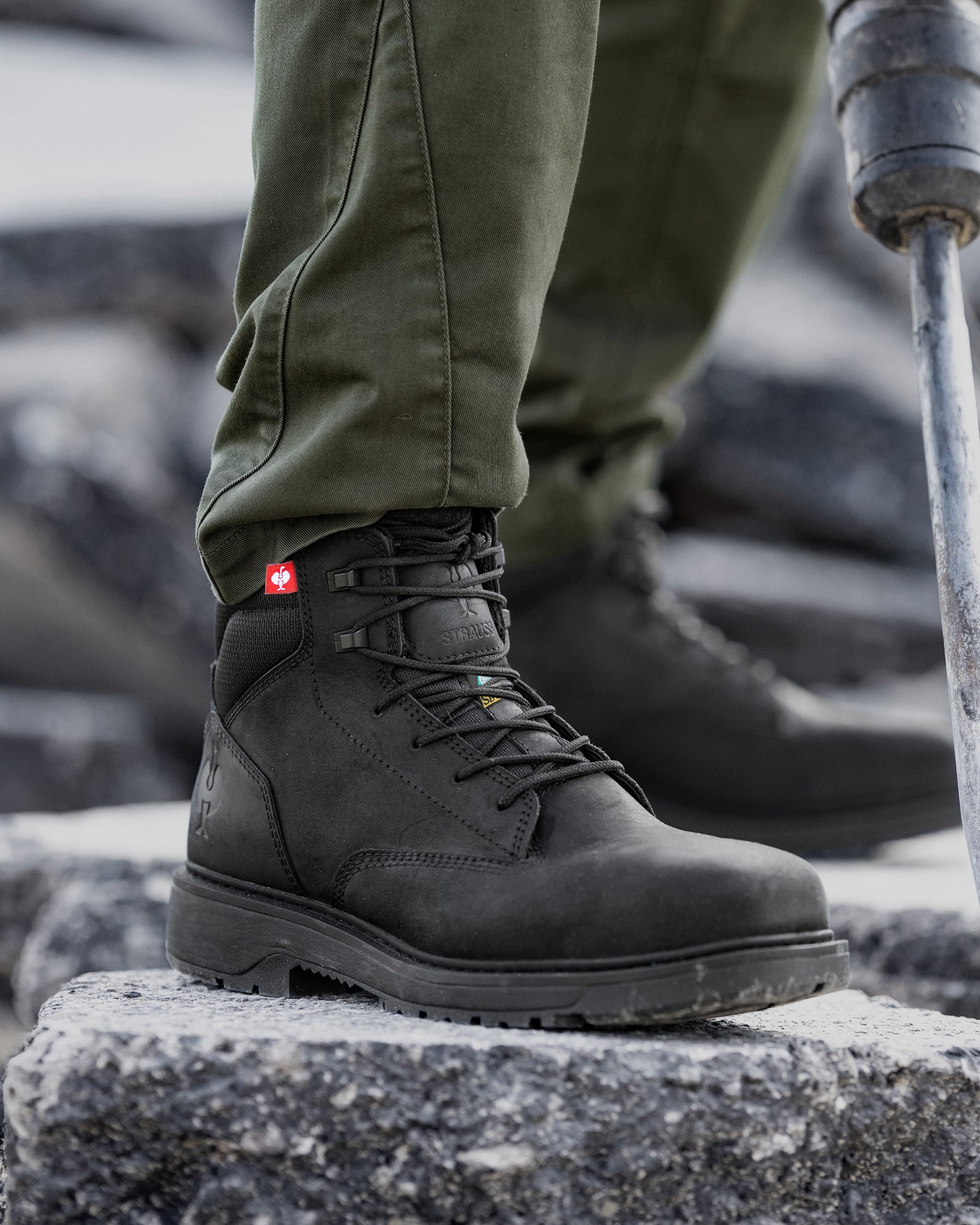 A close-up of a person wearing a black leather hiking boot with olive green pants, standing on a stone surface with a metal trekking pole visible.