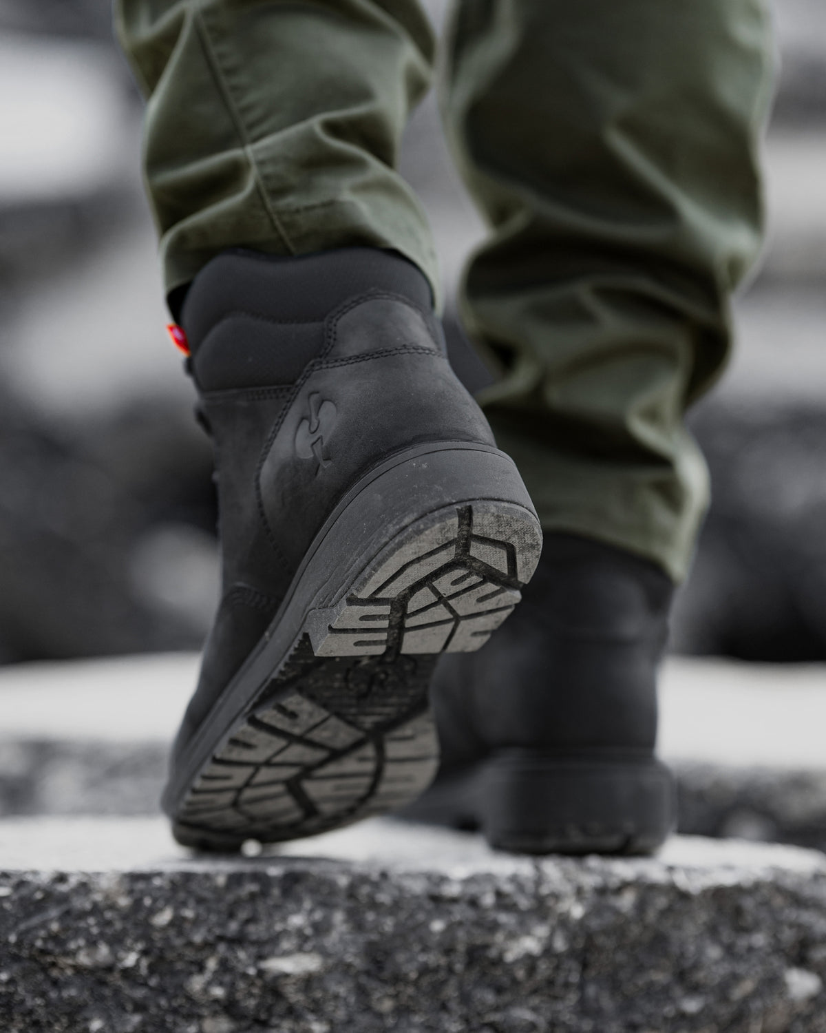 A close-up shot of a person's feet wearing black high-top boots and olive green pants, walking on a wet, dark asphalt surface.