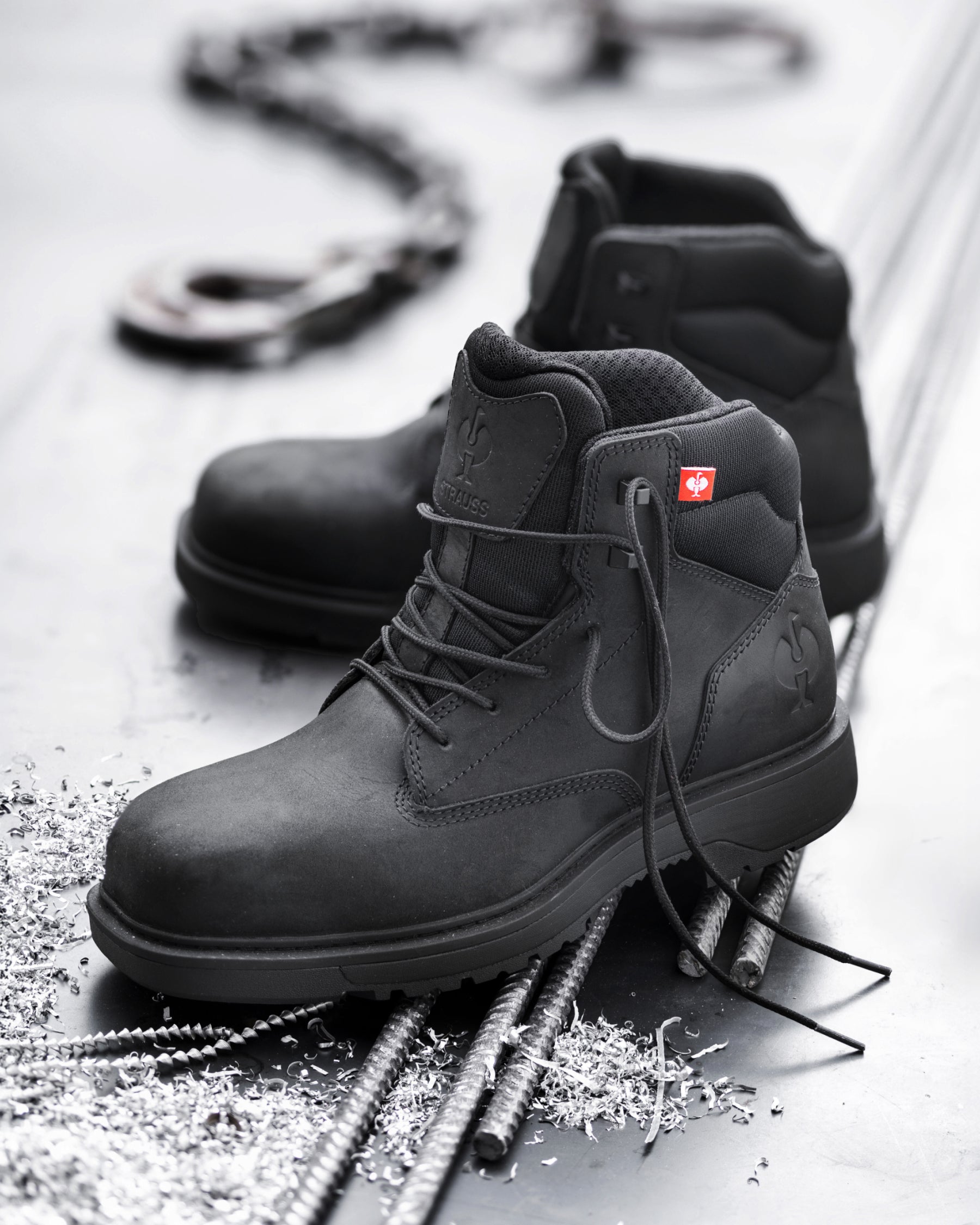 A pair of black high-top work boots arranged on a metallic surface with rebar and small metal shavings. One boot is in sharp focus in the foreground, while the second is slightly blurred behind it.