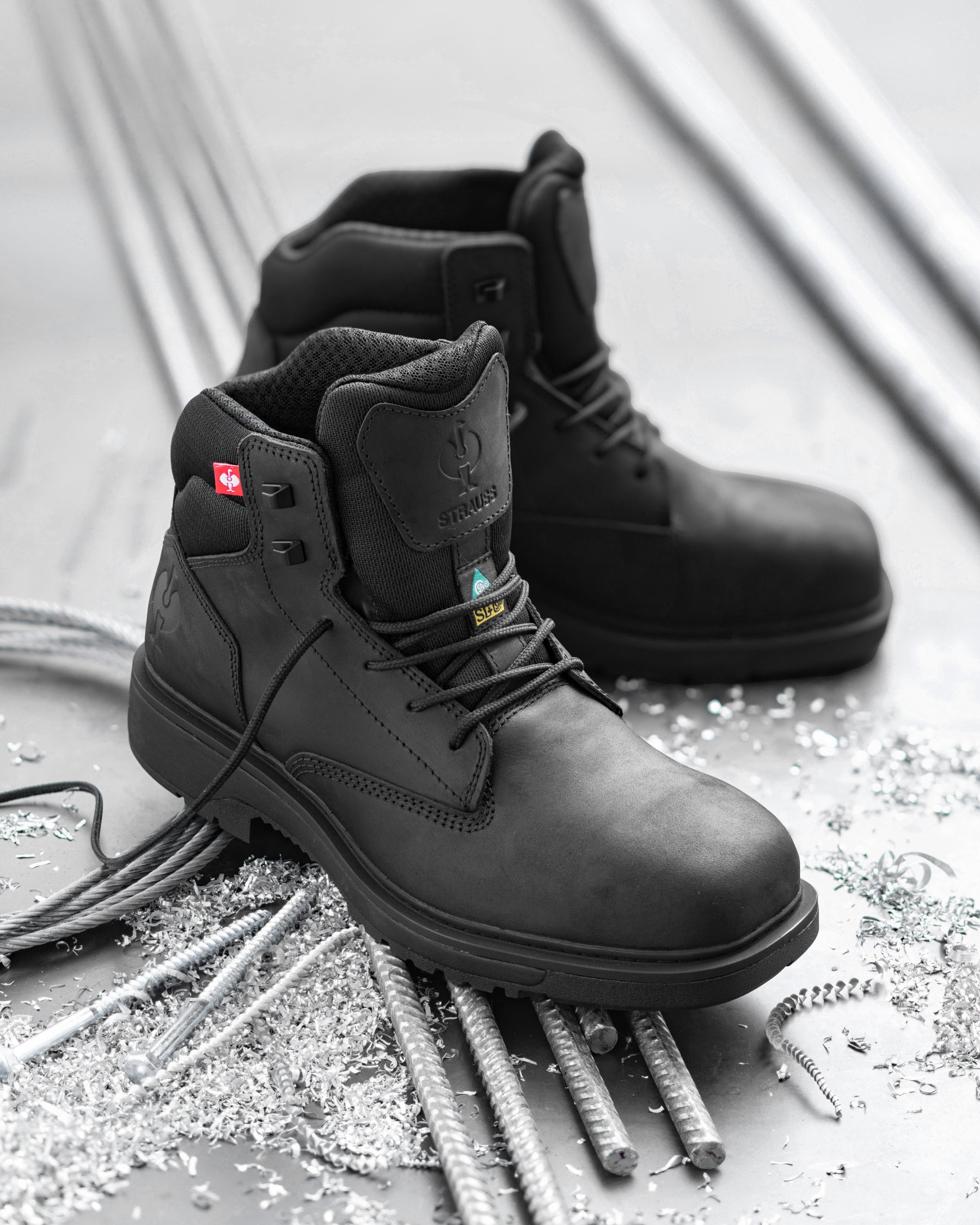 A pair of black industrial safety boots with steel toe protection, displayed on a gray concrete floor surrounded by rebar and metal shavings. The foreground boot is angled to show the side profile, lace-up closure, and heel, while the second boot sits behind it.