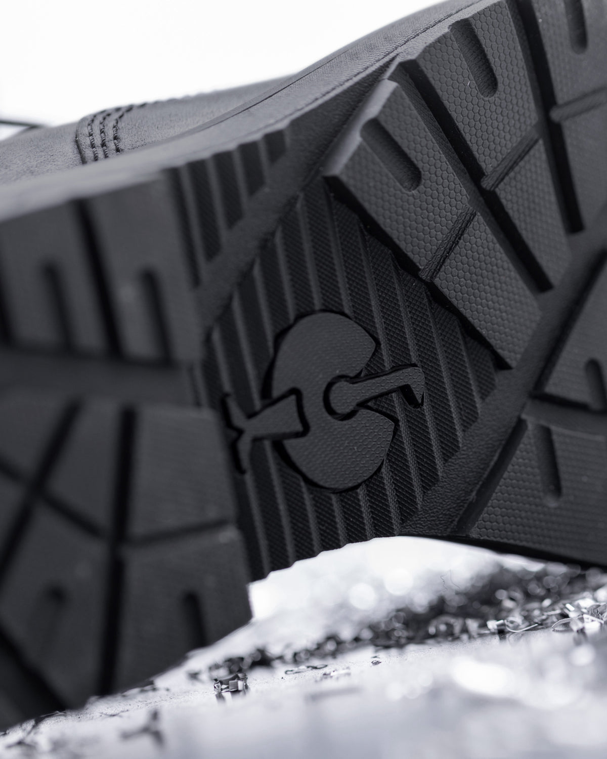 Close-up of the textured black rubber outsole of a shoe, showing a molded logo, photographed over a snowy surface.