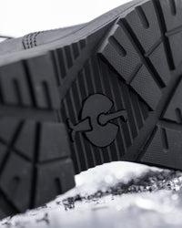 Close-up of the textured black rubber outsole of a shoe, showing a molded logo, photographed over a snowy surface.