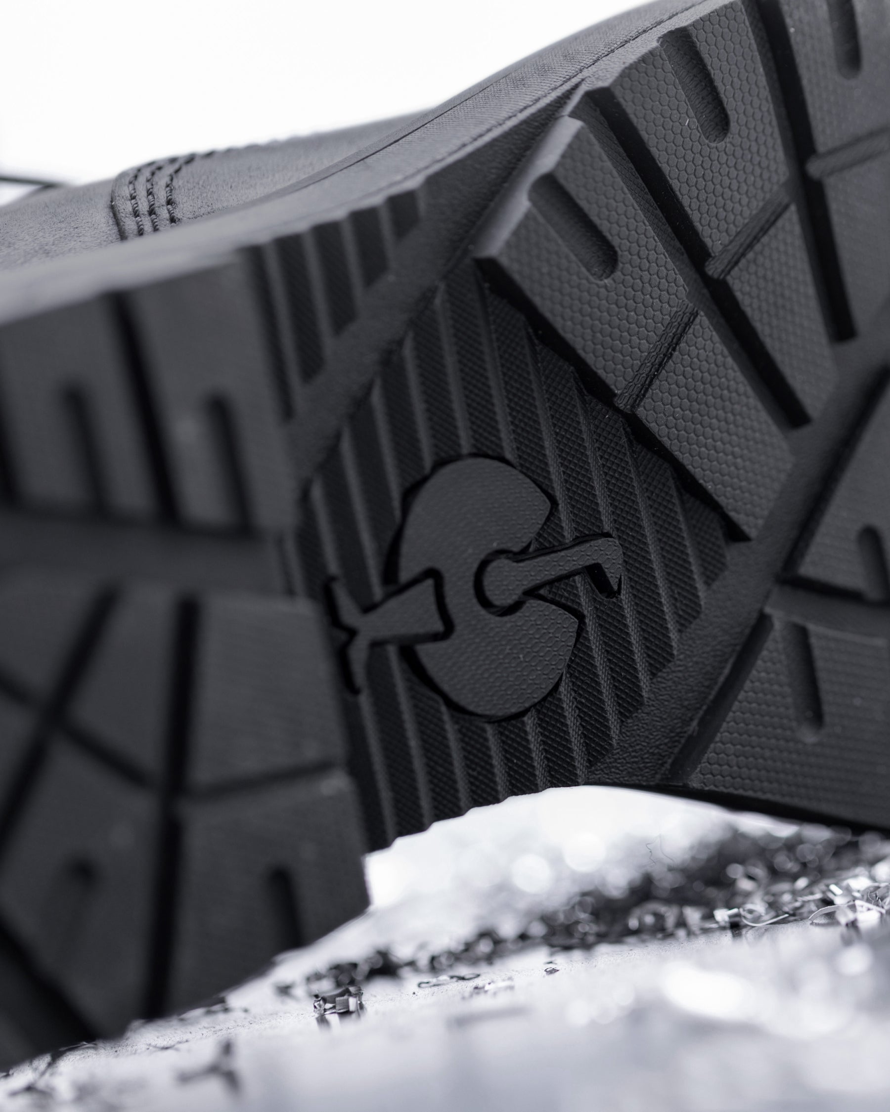 Close-up of the textured black rubber outsole of a shoe, showing a molded logo, photographed over a snowy surface.