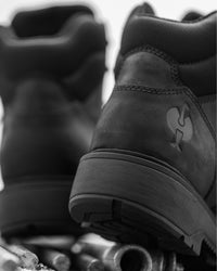 A close-up black-and-white photo of a pair of rugged lace-up boots, with the rear and side of one boot in focus and the other blurred in the background.