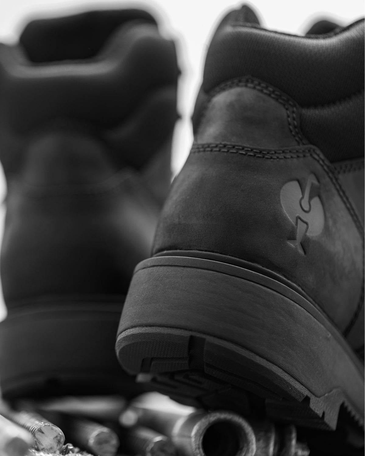 A close-up black-and-white photo of a pair of rugged lace-up boots, with the rear and side of one boot in focus and the other blurred in the background.