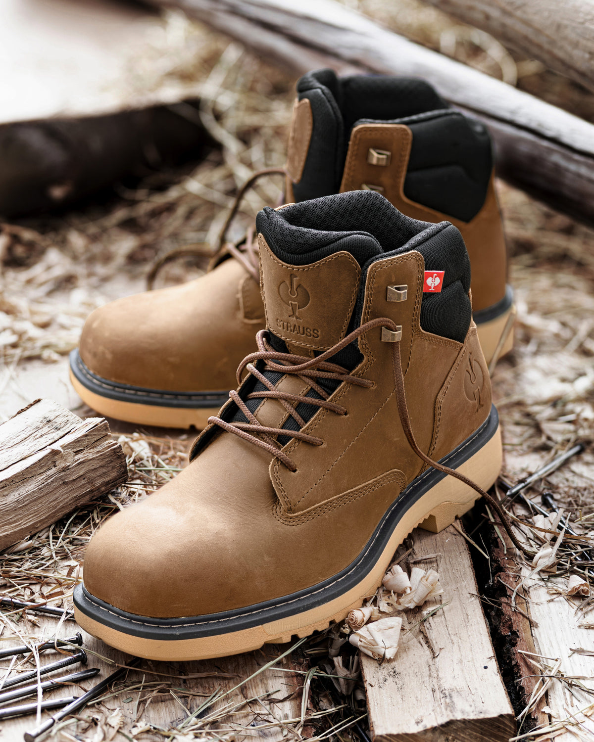 A pair of brown leather lace-up boots with black ankle lining and beige soles, placed on a rustic bed of wood and dry grass. A small Swiss flag patch is visible on the collar.