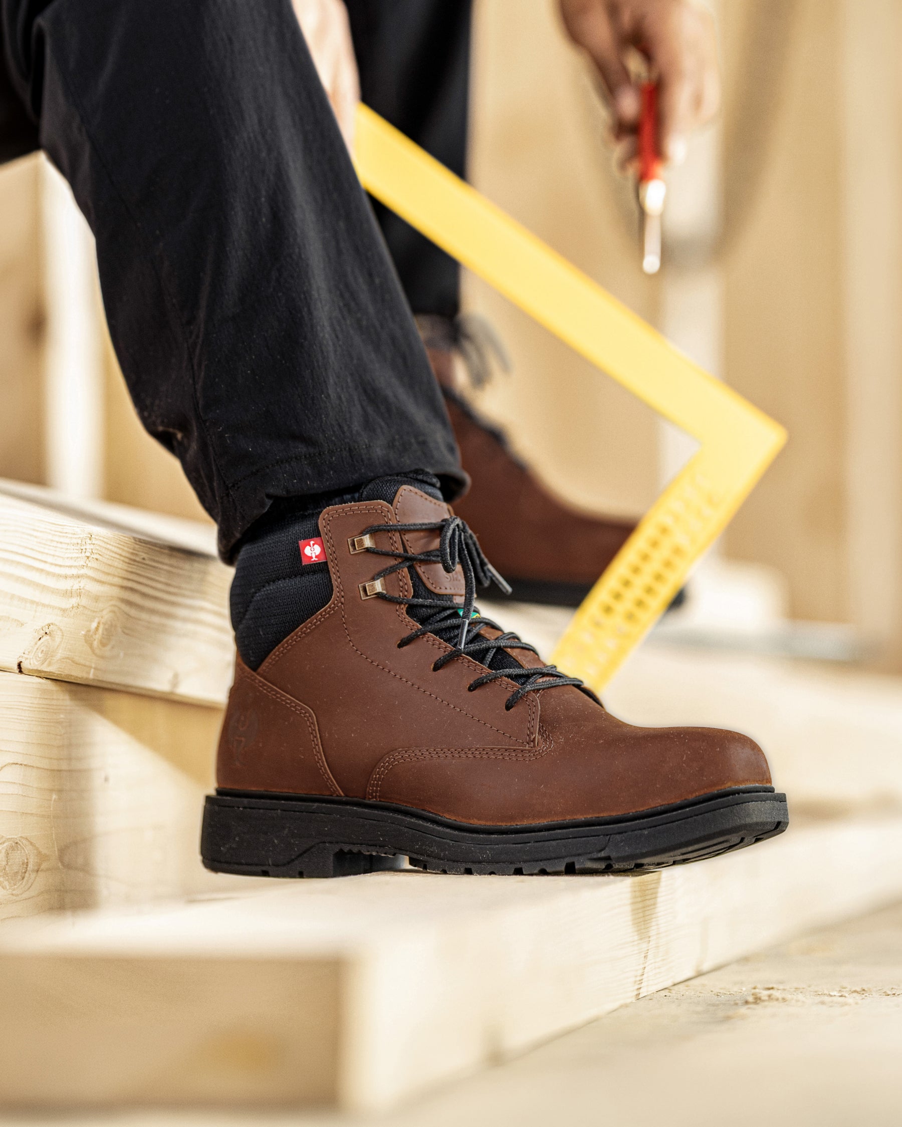 A close-up of a person wearing a brown leather work boot with black laces and a black sock, standing on wooden construction steps. A yellow square ruler and a hand holding a screwdriver are visible in the background.