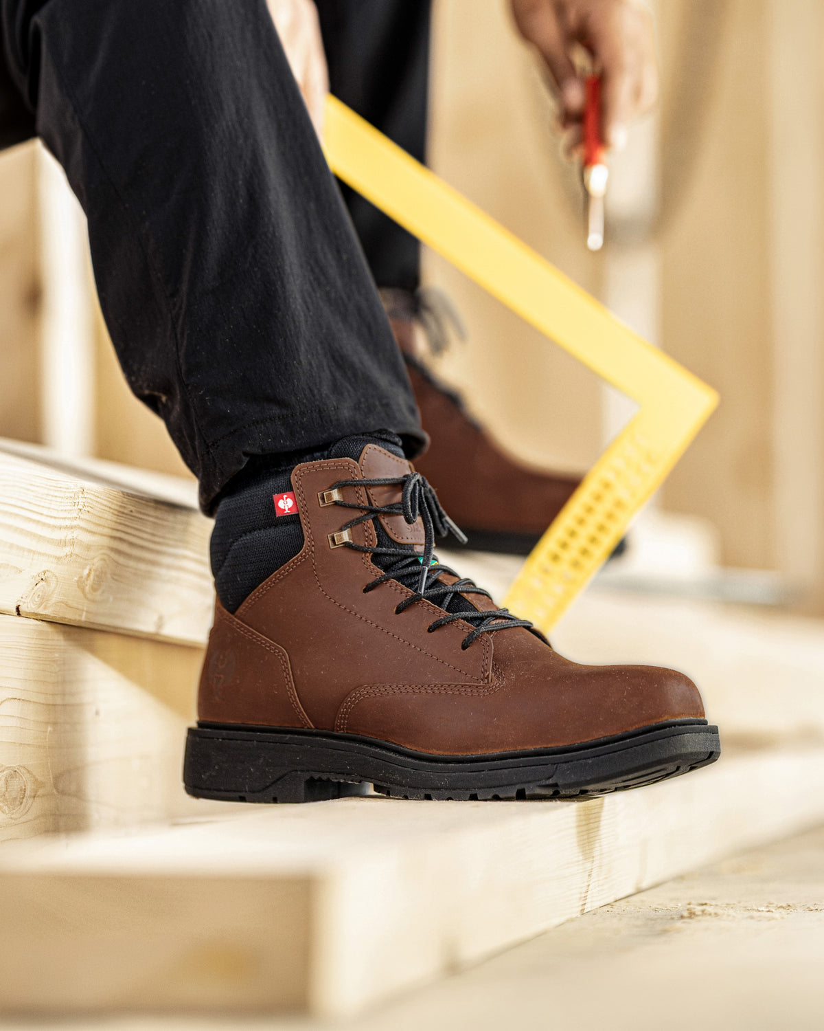 A close-up of a person wearing a brown leather work boot with black laces and a black sock, standing on wooden construction steps. A yellow square ruler and a hand holding a screwdriver are visible in the background.