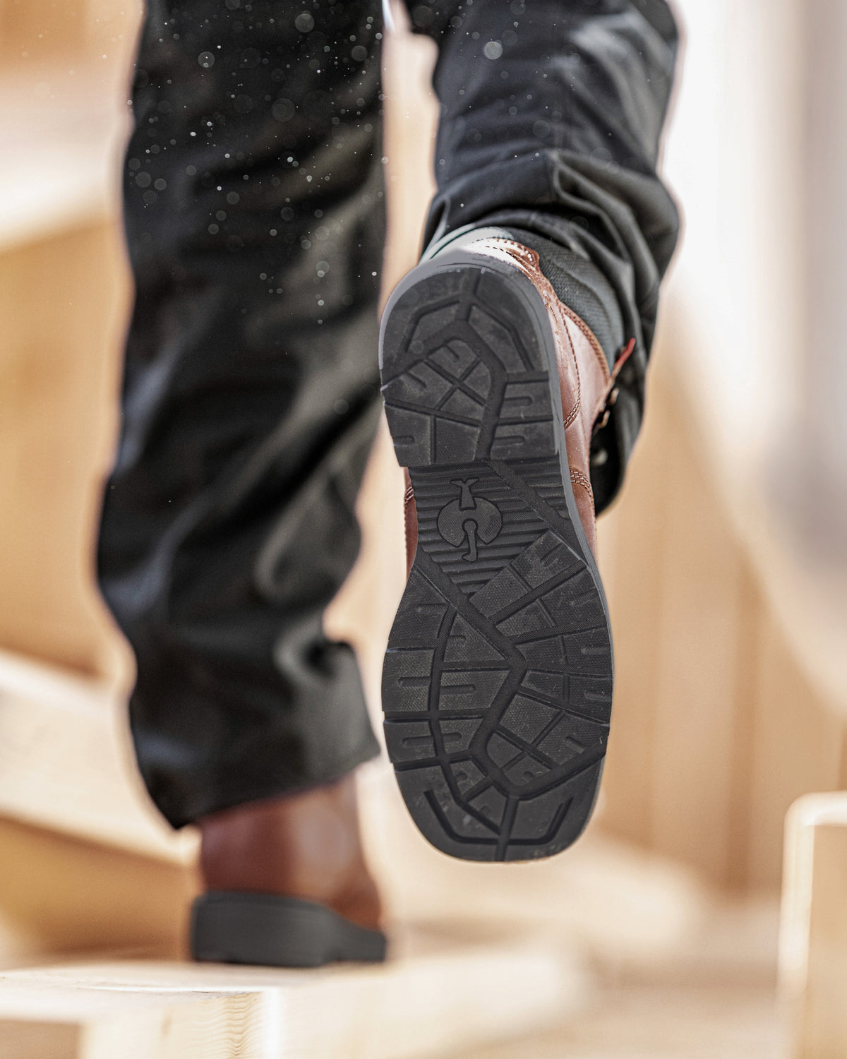 A close-up action shot of a person's leg lifting a brown leather work boot off a wooden step, showing the detailed tread pattern of the black rubber outsole.