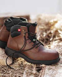 A pair of brown leather work boots with black soles and laces, styled on a rustic background of straw and wood.