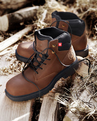 A pair of brown leather lace-up boots with black soles and black lining, placed on weathered wooden planks outdoors with dry grass and logs in the background.