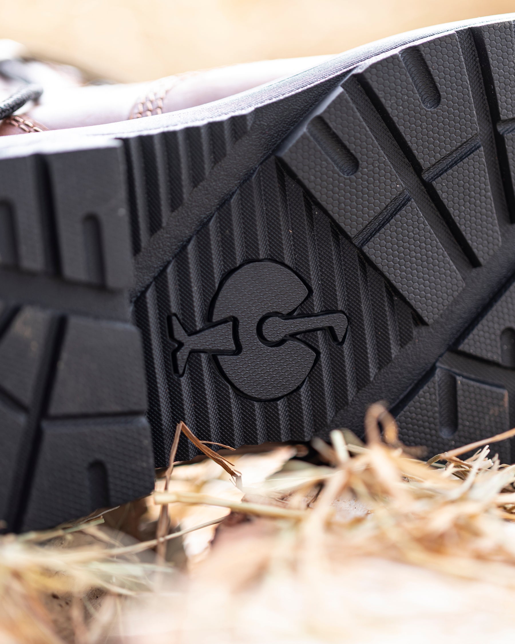 Close-up of the black rubber outsole of a boot, showing a molded logo and tread pattern, resting on dry straw with shallow depth of field.