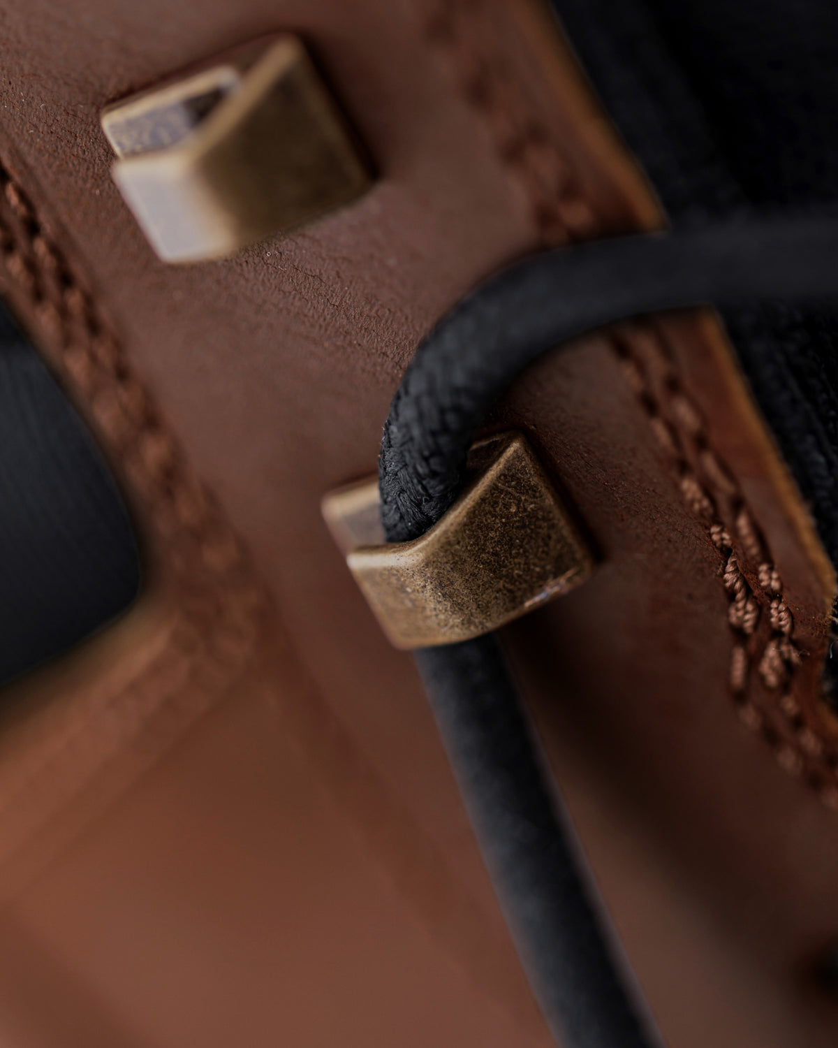 Close-up detail of a brown leather boot with black laces threaded through square brass eyelets.
