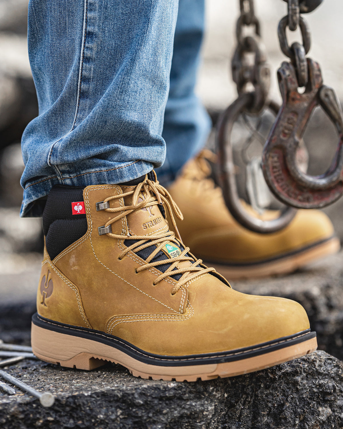 A close-up of a person wearing a tan suede work boot with blue jeans, standing on a rough concrete surface next to hanging metal chains and hooks. The boot has tan laces, a black collar, and a tan rubber outsole.