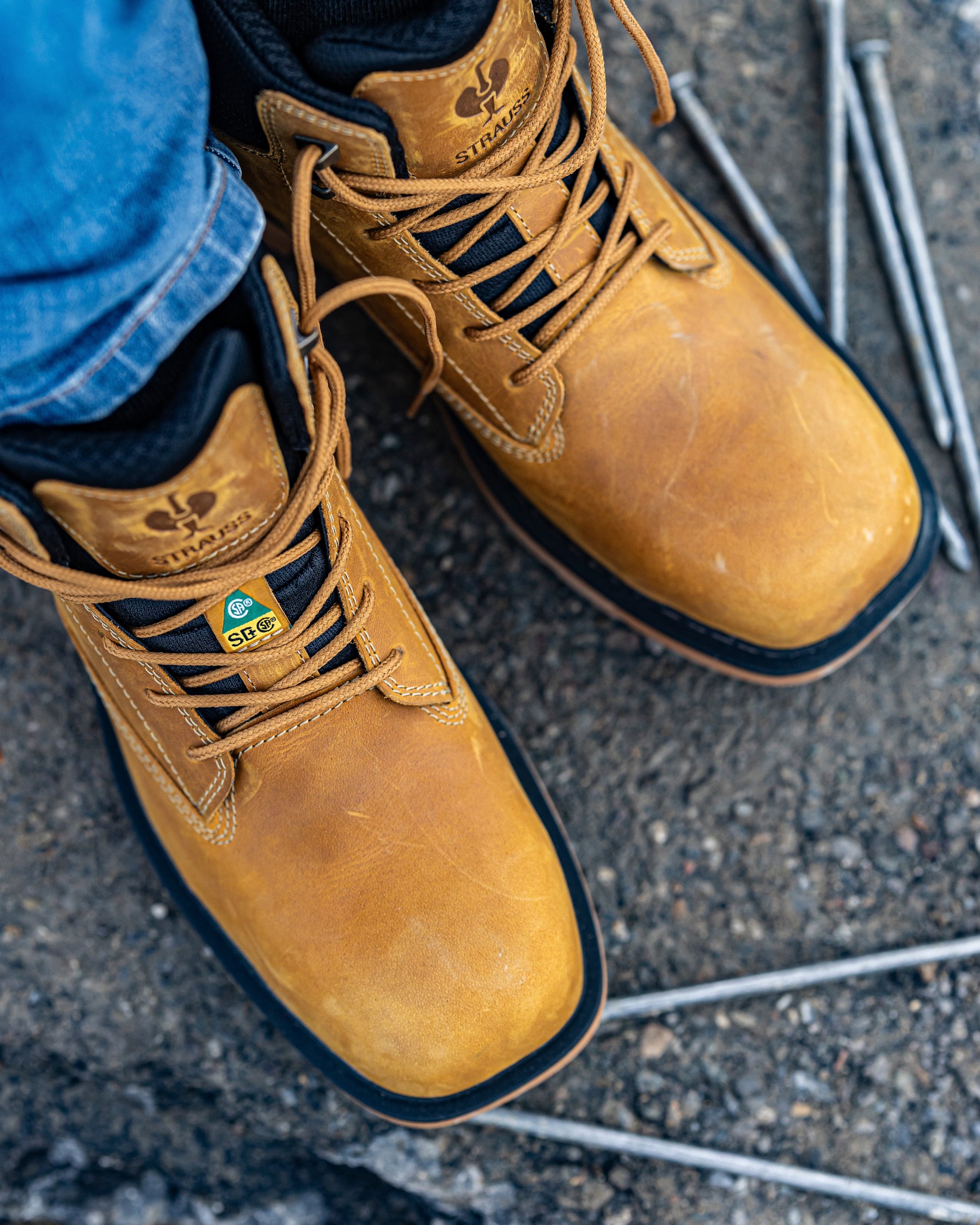 A close-up of a person wearing tan work boots with blue jeans, standing on a rough concrete surface with metal nails nearby.