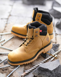 A pair of tan work boots with black ankle lining and a small Swiss flag detail on the tongue, displayed on a construction site with concrete debris and metal screws.