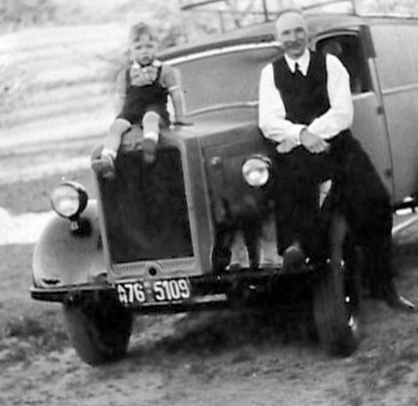 A young boy sits on the hood of a vintage car while an older man leans against the front. Both are dressed formally. The photo is black and white and has a nostalgic, old-fashioned feel.