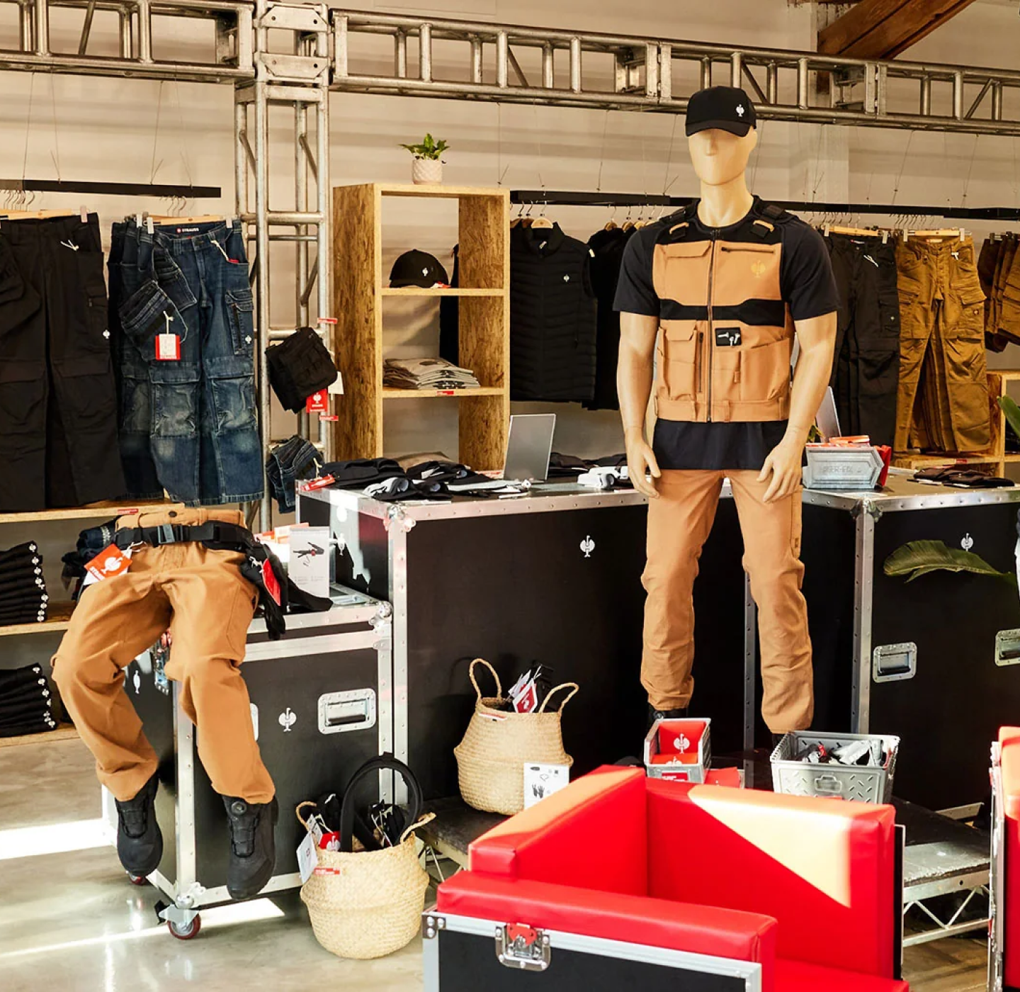 A mannequin in brown workwear and a black hat stands beside equipment cases in a clothing store, with pants, shirts, baskets, and other retail items displayed around the industrial-style space.