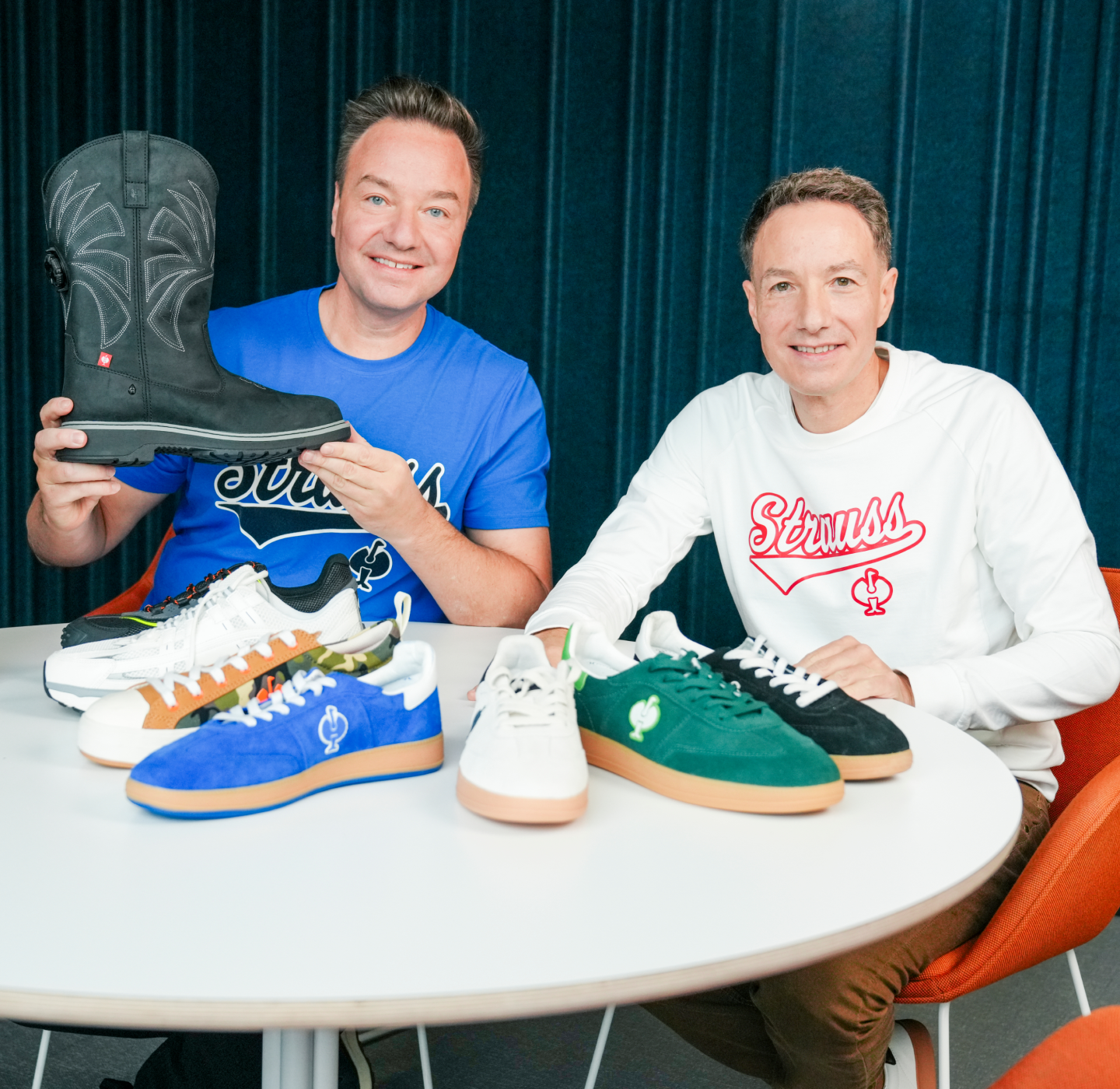 Two men sit at a table displaying five pairs of colorful sneakers and one black boot. Both are smiling; one wears a blue shirt, the other a white shirt. Blue curtains form the background.
