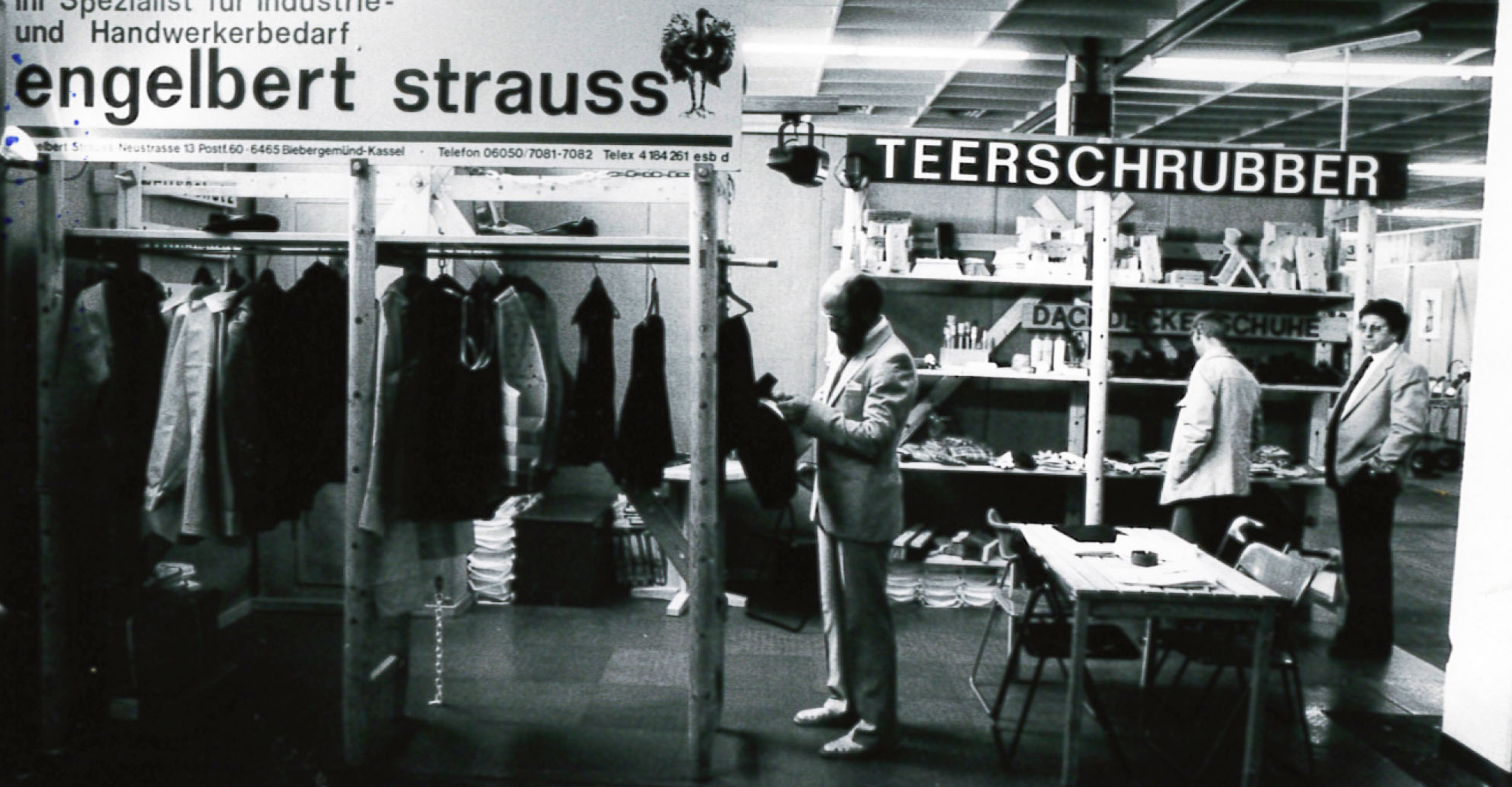 Black-and-white photo of men in suits at a trade fair booth displaying workwear and tools, with signs reading engelbert strauss and TEERSCHRUBBER above the stalls.
