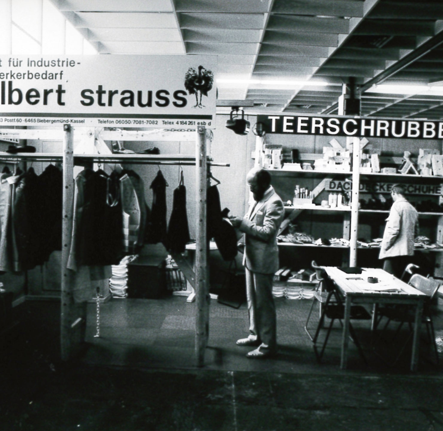 A man in a suit examines a jacket at a clothing booth in an indoor market or trade show. Signs above the booths read Albert Strauss and Teerschrubb. Tables and racks display garments and other items.