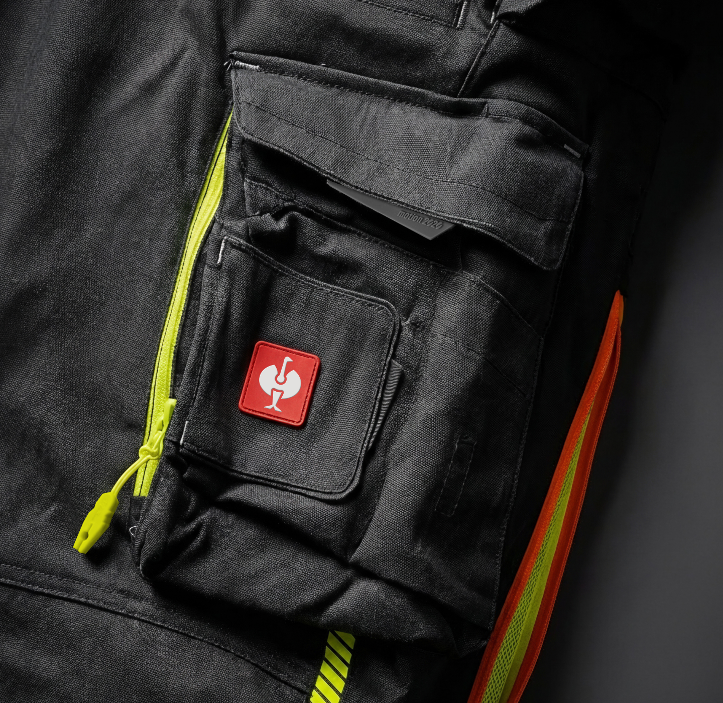 A close-up of a black utility garment with bright yellow zippers, orange accents, and multiple pockets. One pocket features a red patch with a white fragile glass symbol.