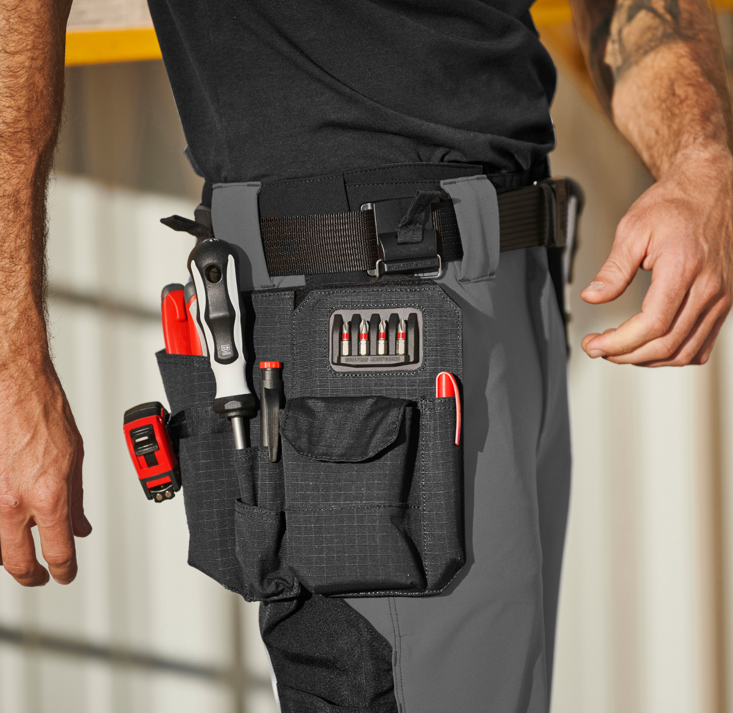 A person wearing gray work pants and a black belt with a tool pouch containing a screwdriver, pliers, red pen, utility knife, small tools, and bits. Only the persons waist and hand are visible.
