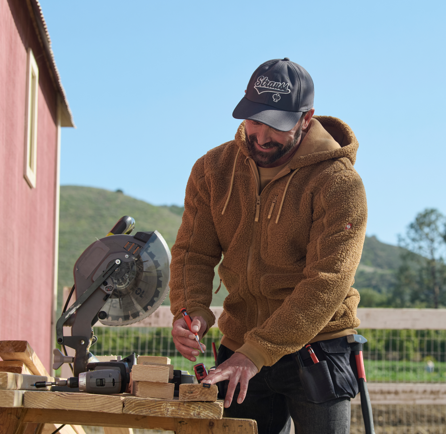 A man wearing a brown hoodie and black cap measures a wooden board with a tape measure and pencil at an outdoor workbench, with a miter saw and tools nearby. A red building and hills are in the background.