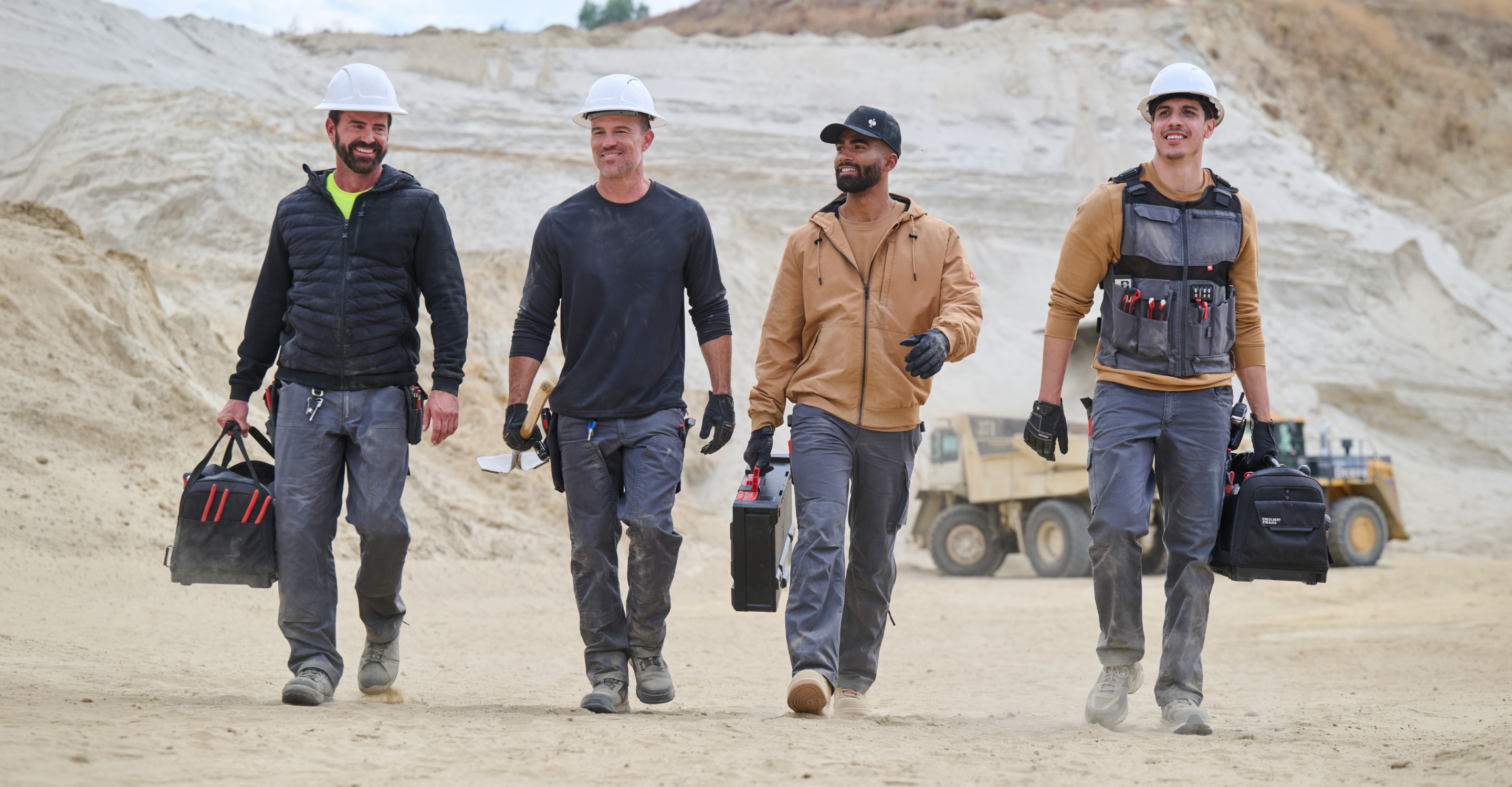 Four construction workers wearing hard hats, gloves, and work gear walk side by side in a sandy worksite, carrying tool bags. Construction vehicles and equipment are visible in the background.