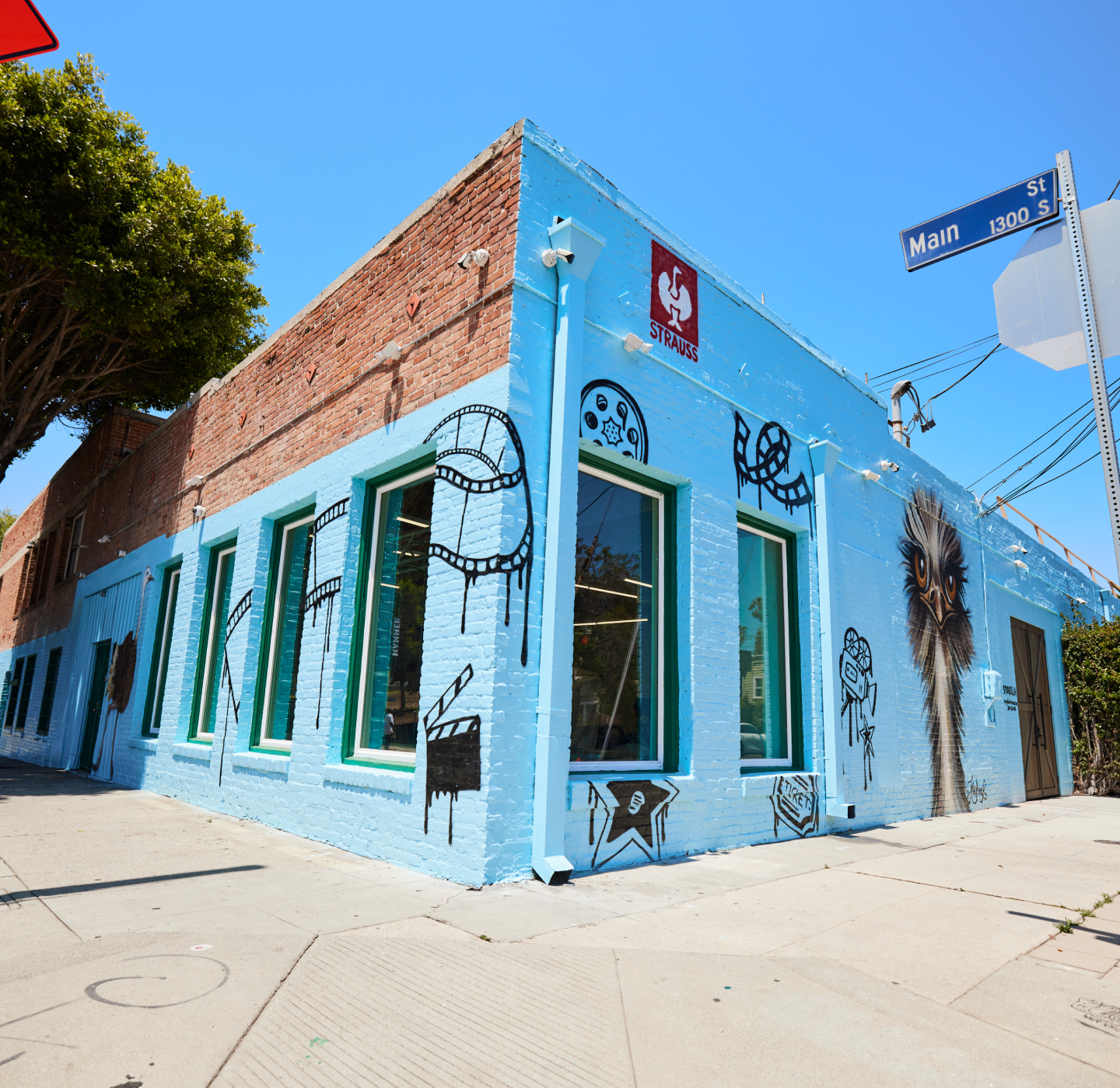 A blue-brick building on a sunny street corner, decorated with black film-themed murals, including camera reels and a directors chair, with a Main St sign visible.