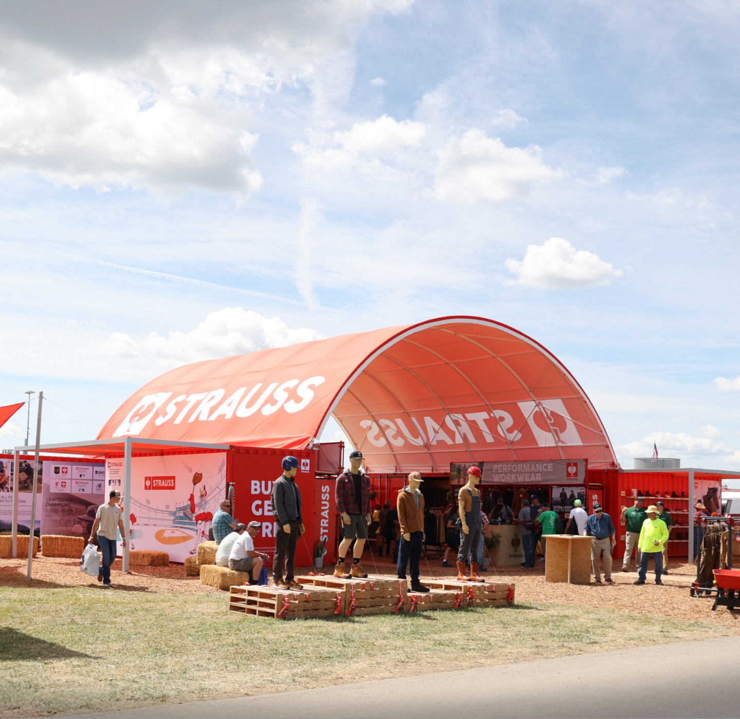 People gather outside a large red tent labeled STRAUSS at an outdoor event. Some are sitting on hay bales, while others stand and walk around under a partly cloudy sky.