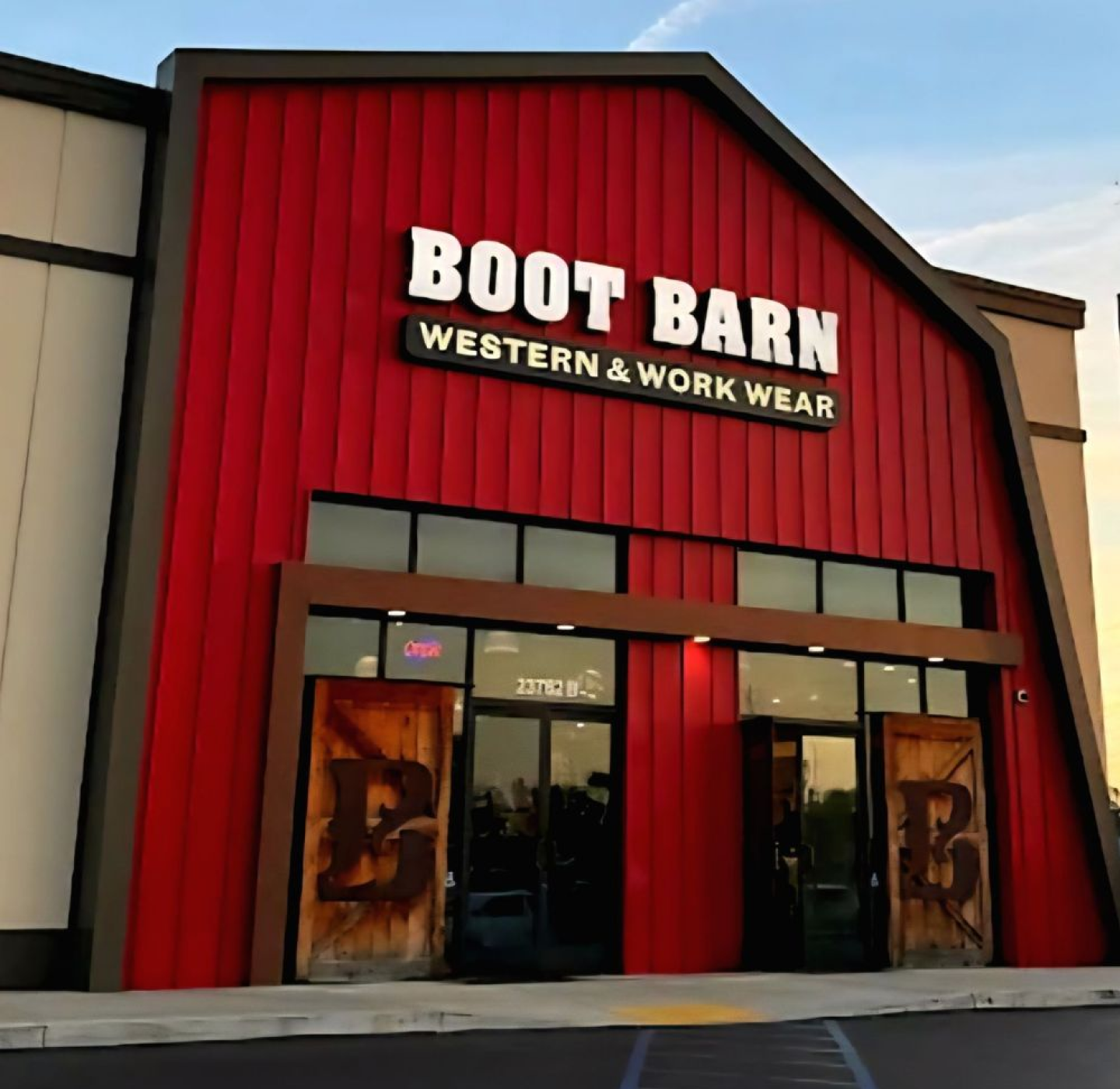 A red building with a large sign reading BOOT BARN WESTERN & WORK WEAR above double wooden doors. The store appears open, and the exterior has a western-style design.