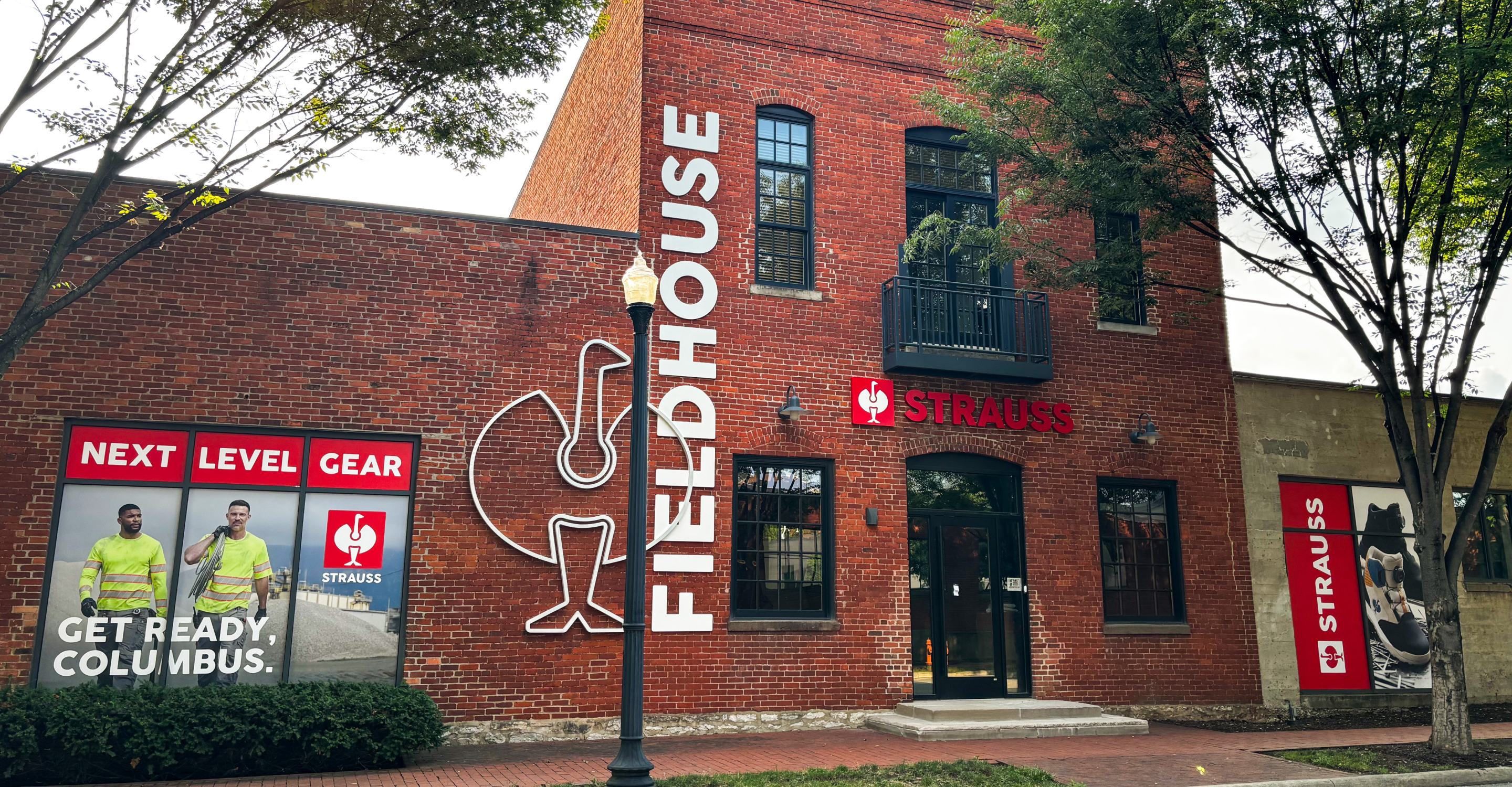 A red brick building labeled Fieldhouse and Strauss with large windows, signage, and two people in safety gear on a poster that says Next Level Gear. Get Ready, Columbus. Trees frame the scene.