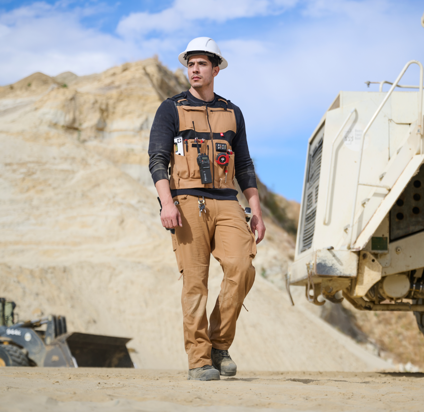 A construction worker in safety gear stands on a sandy site near large machinery, with rocky hills and blue sky in the background.