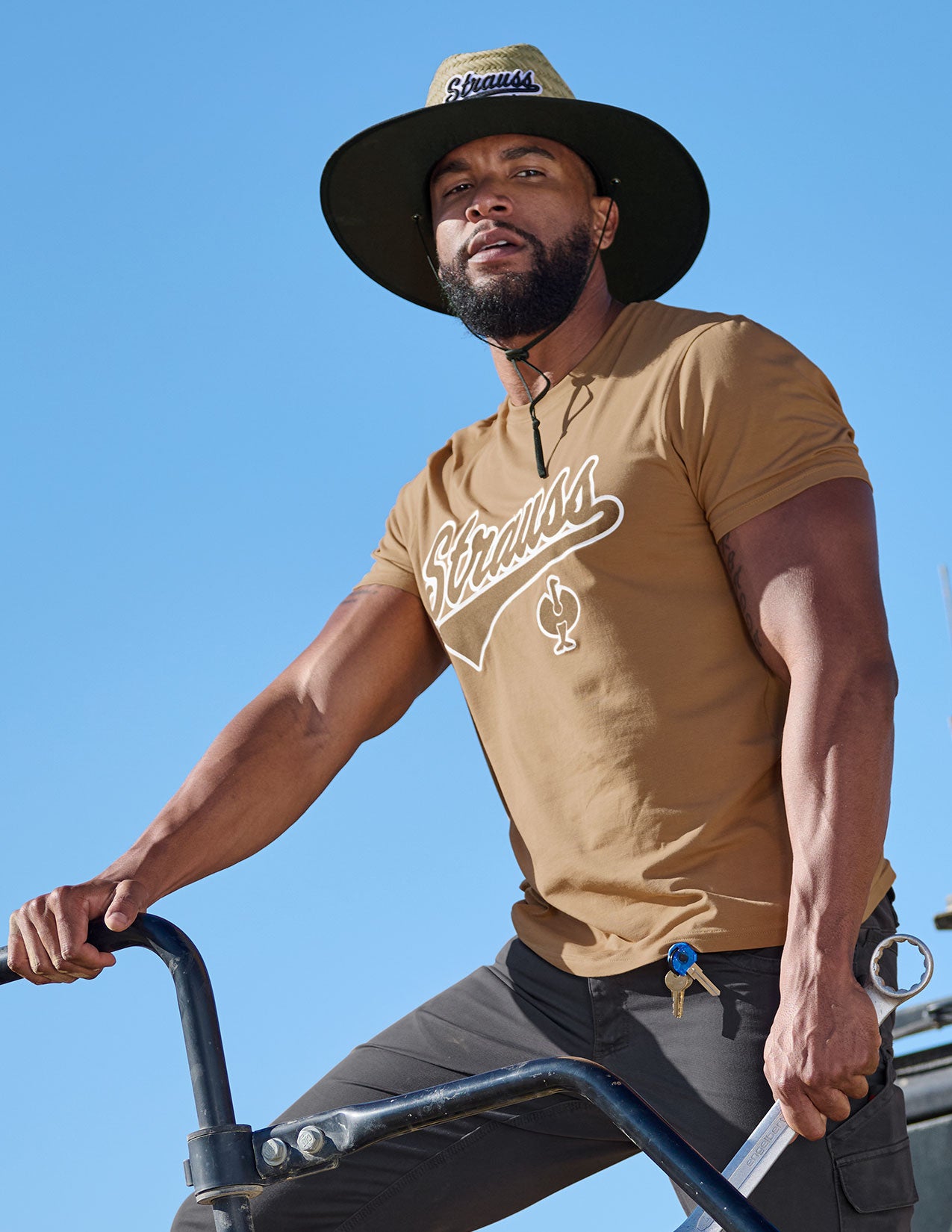 Man wearing a brown t-shirt with a logo and a straw hat, standing outdoors against a clear blue sky.