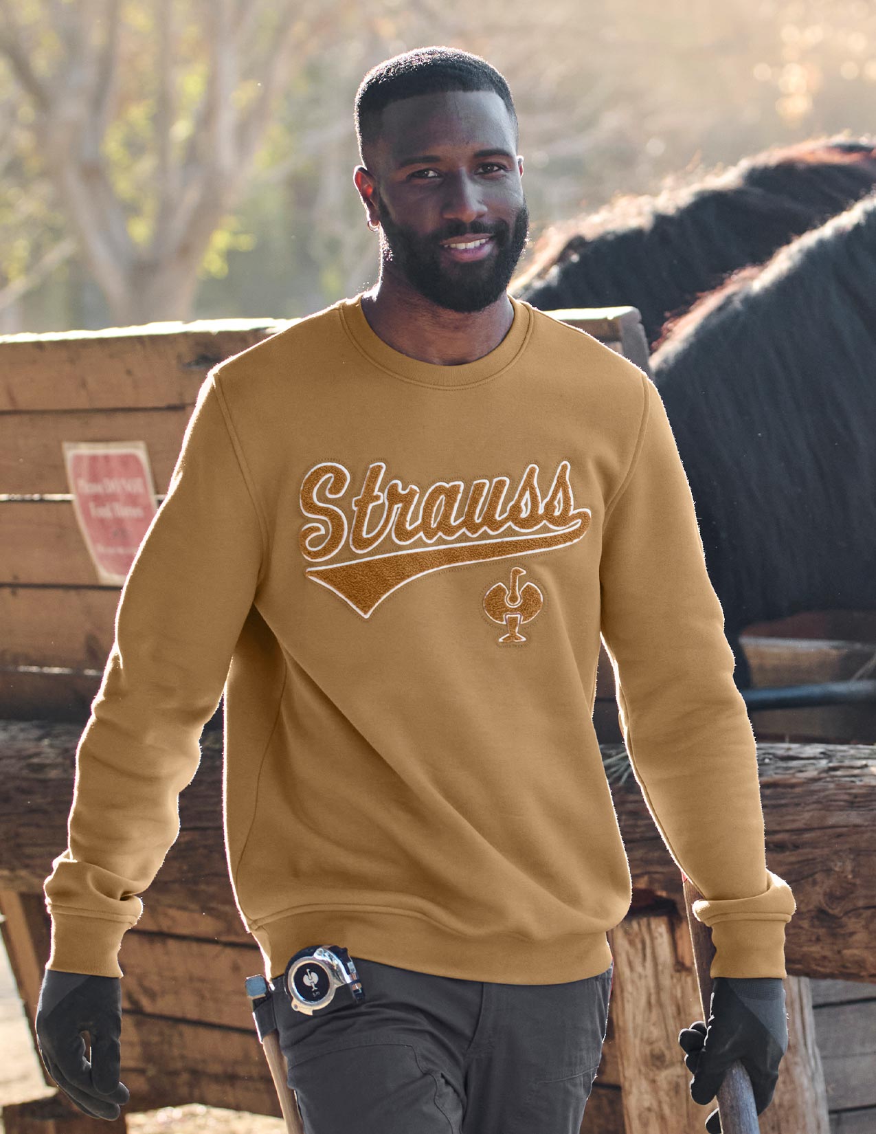 Man wearing a brown 'Strauss' sweatshirt standing outdoors with wooden structures and a horse in the background.