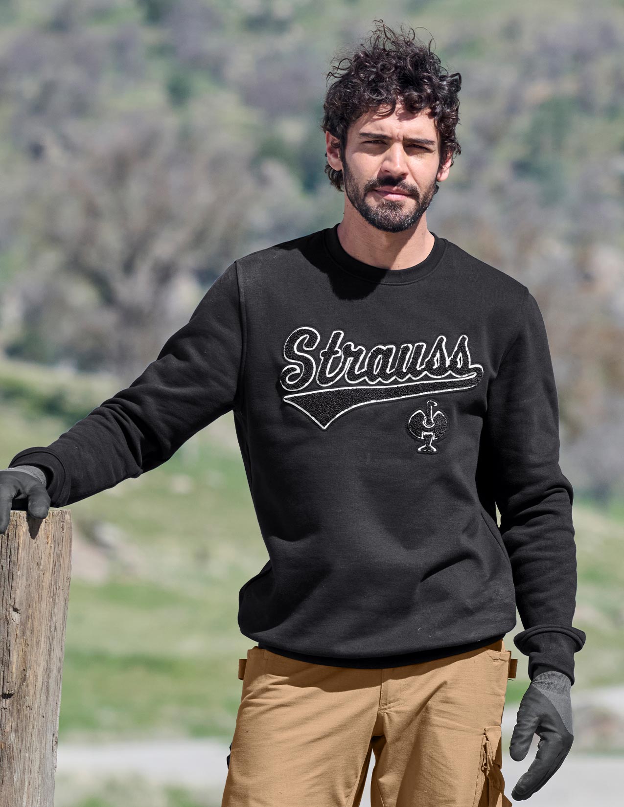 Man wearing a black 'Strauss' sweatshirt outdoors with greenery in the background