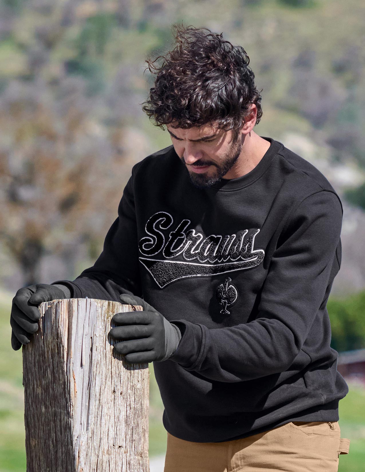 Man wearing a black sweatshirt with a logo, holding a wooden post outdoors.