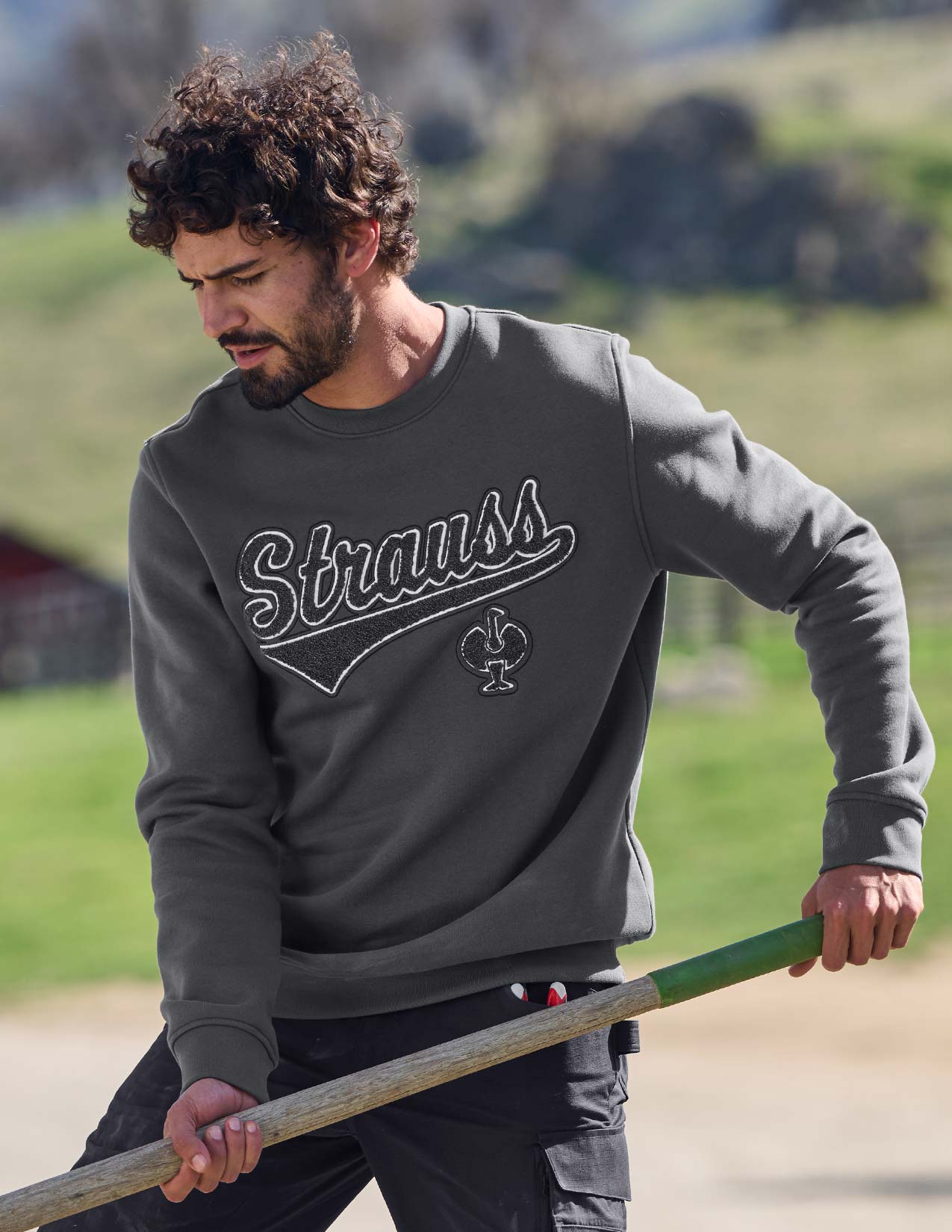 Man wearing a gray 'Strauss' sweatshirt holding a green and brown stick outdoors.