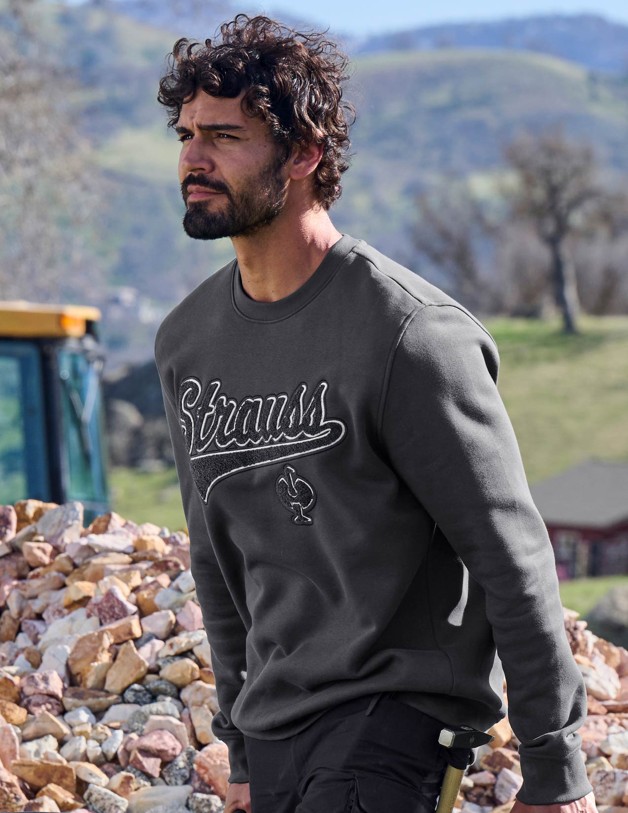 Man wearing a dark gray 'Strauss' sweatshirt standing outdoors with mountains in the background