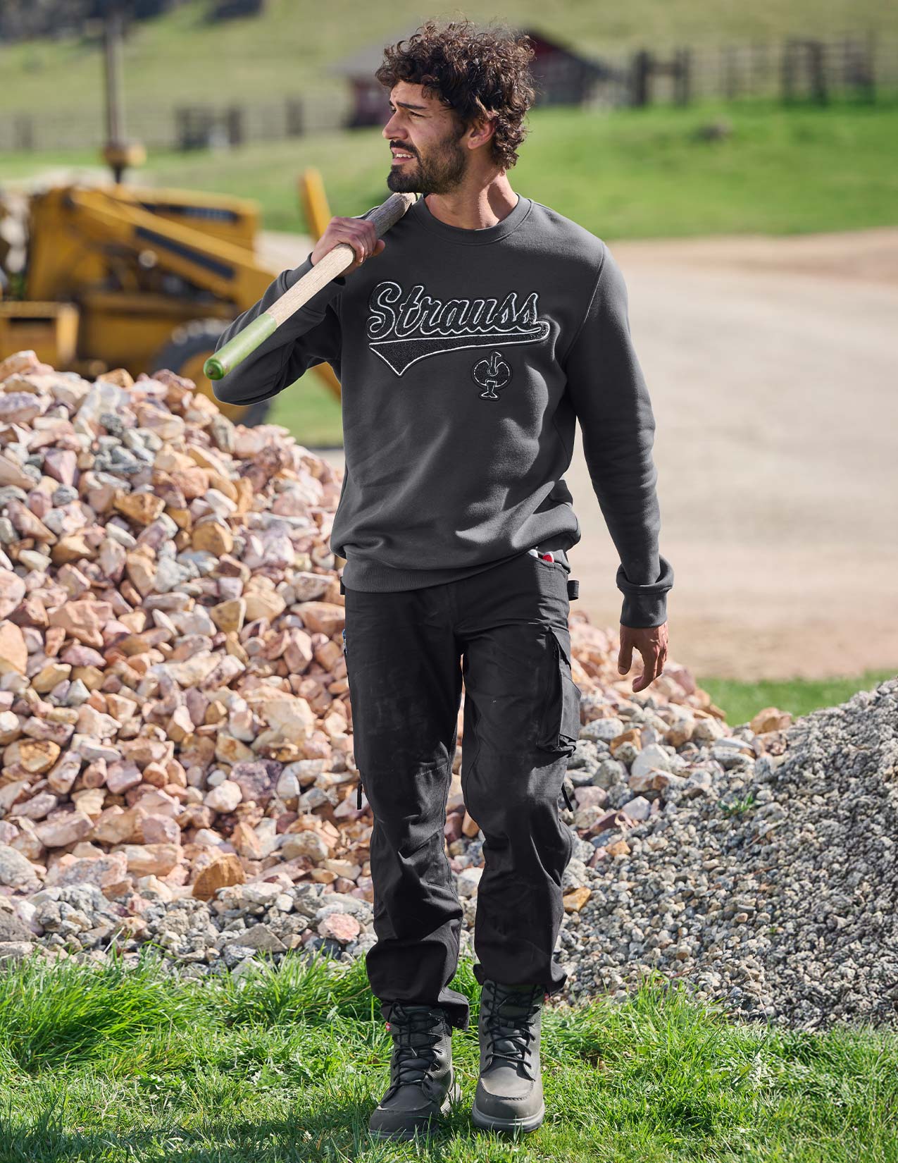 Man wearing a 'Strauss' sweatshirt carrying a shovel outdoors.