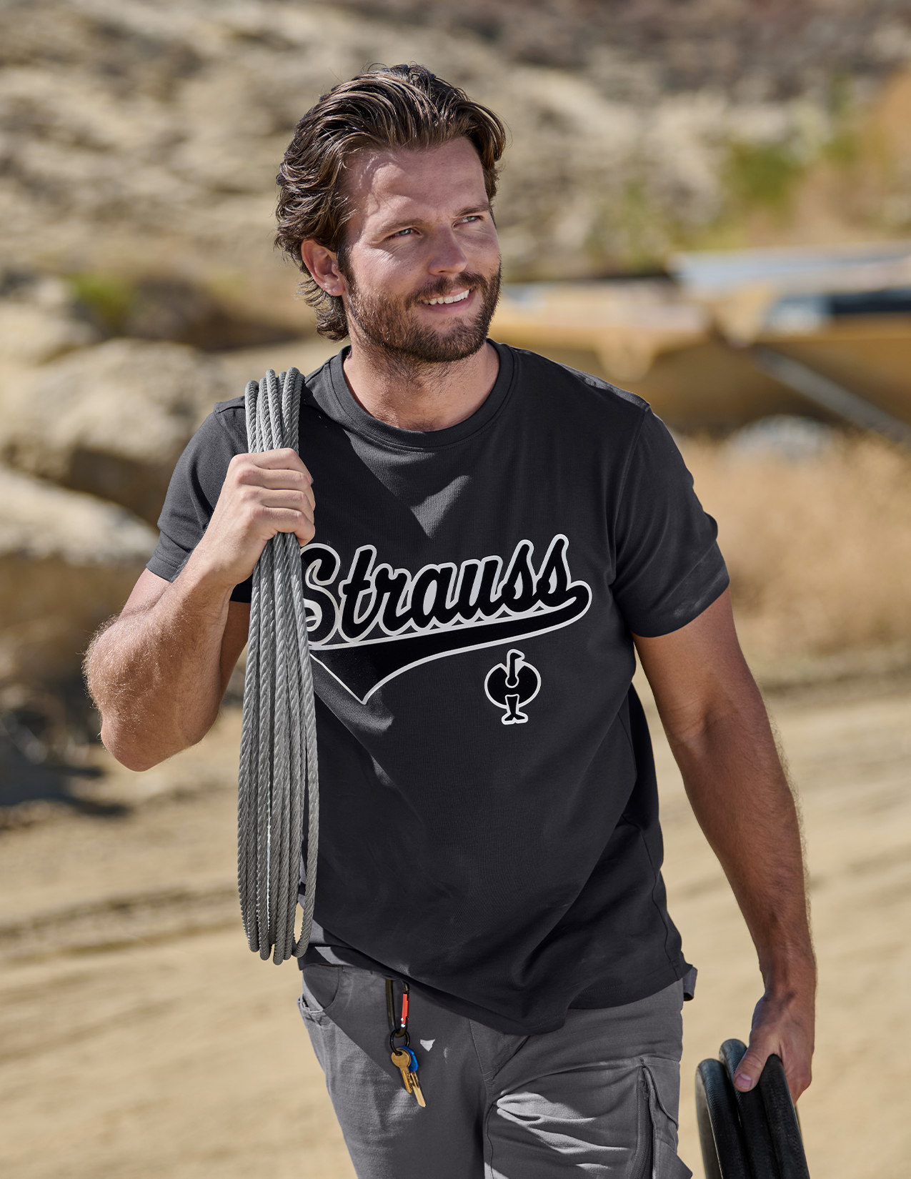Man wearing a black t-shirt with 'Strauss' logo, holding climbing gear in a natural setting.