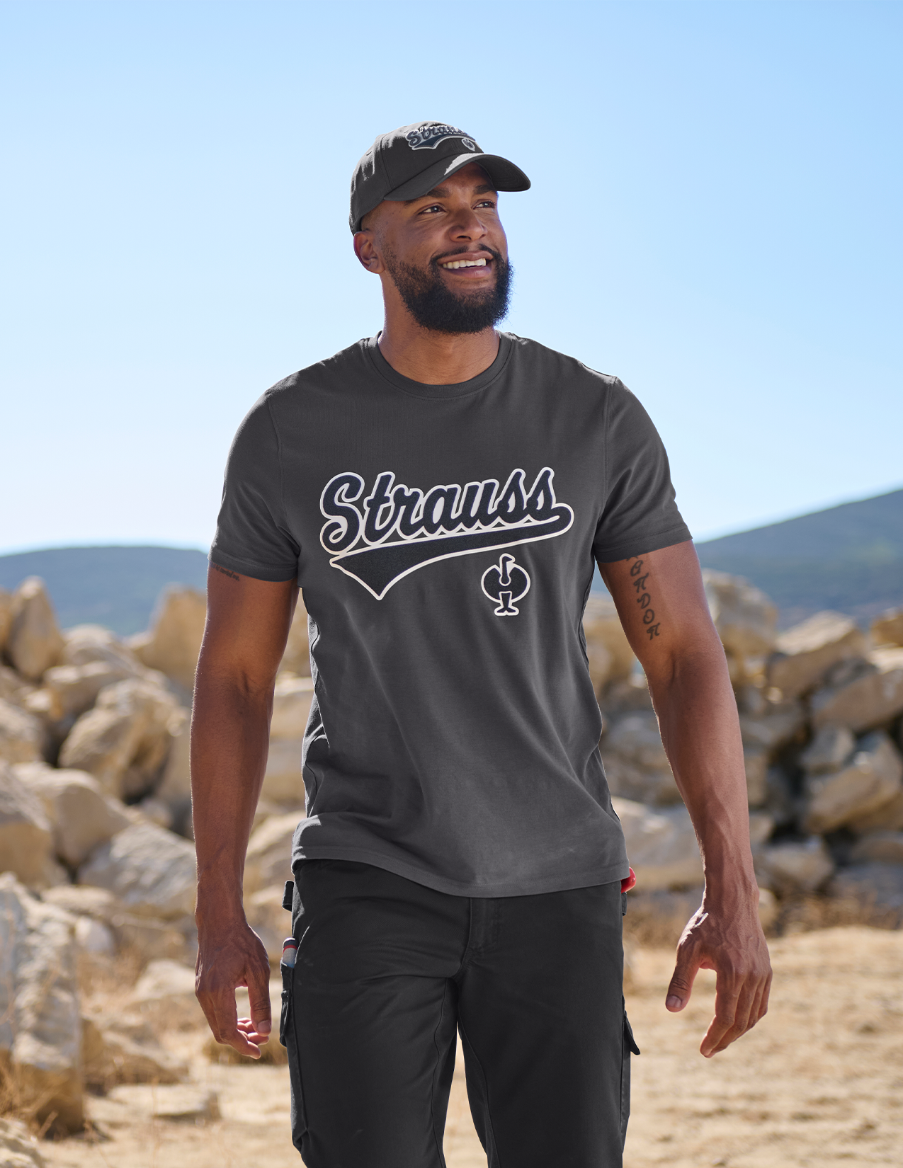 Man wearing a gray 'Strauss' t-shirt standing outdoors with rocky landscape and clear sky.