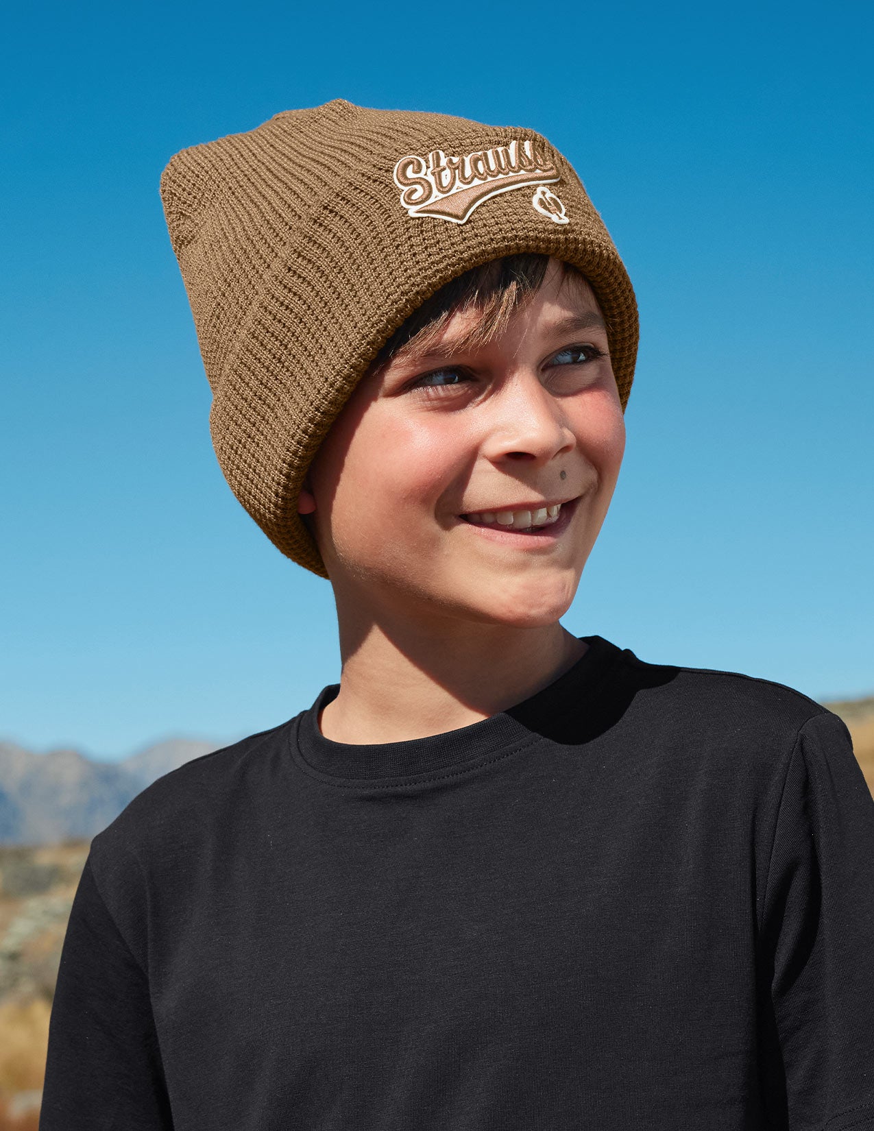 Child wearing a brown knit beanie with a logo against a clear blue sky