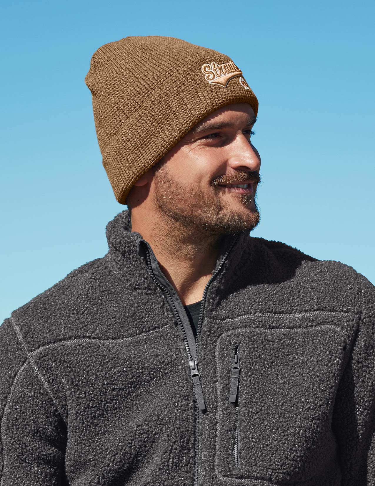 Man wearing a brown knit beanie and dark gray fleece jacket against a clear blue sky.