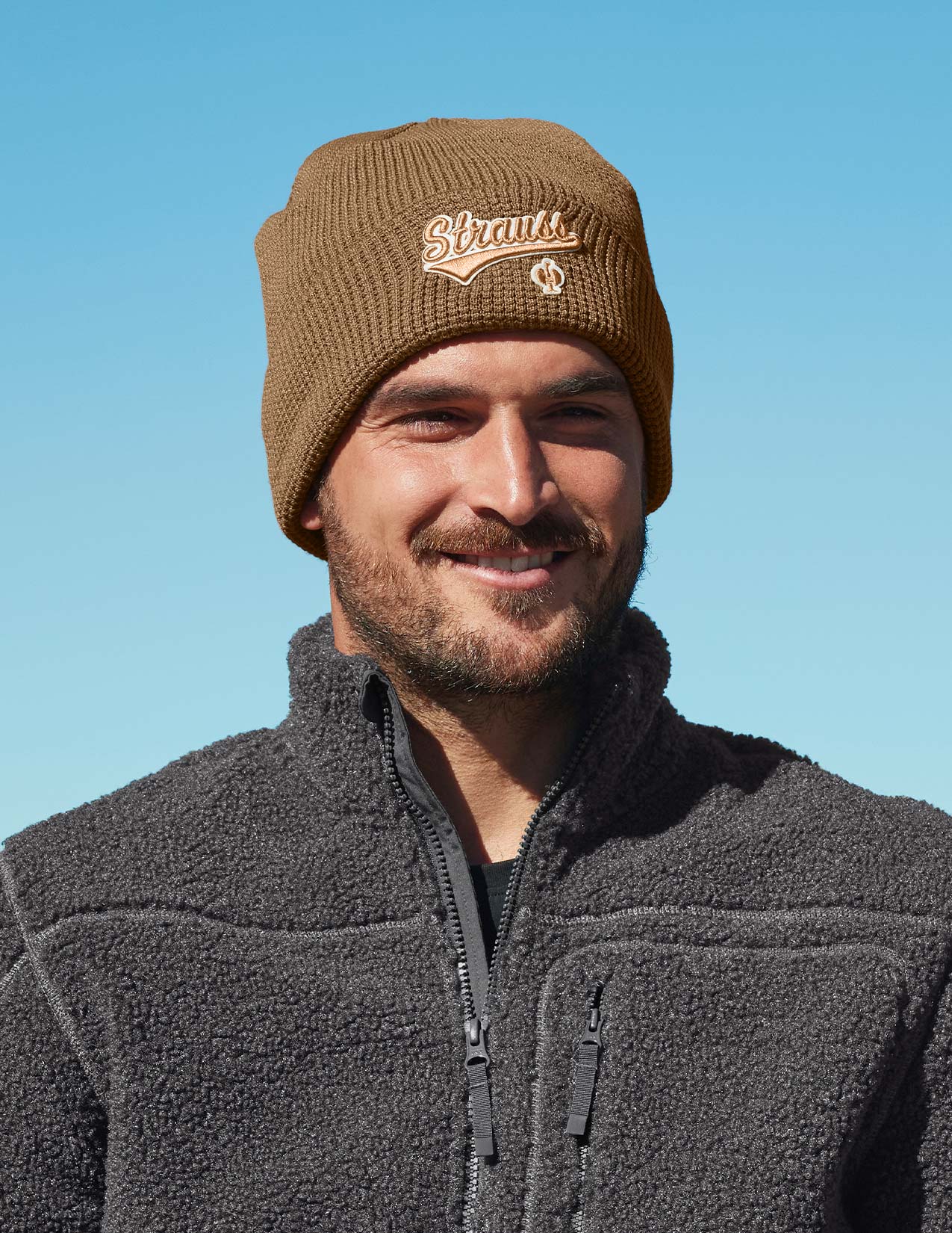 Man wearing a brown knit beanie with branding against a clear blue sky
